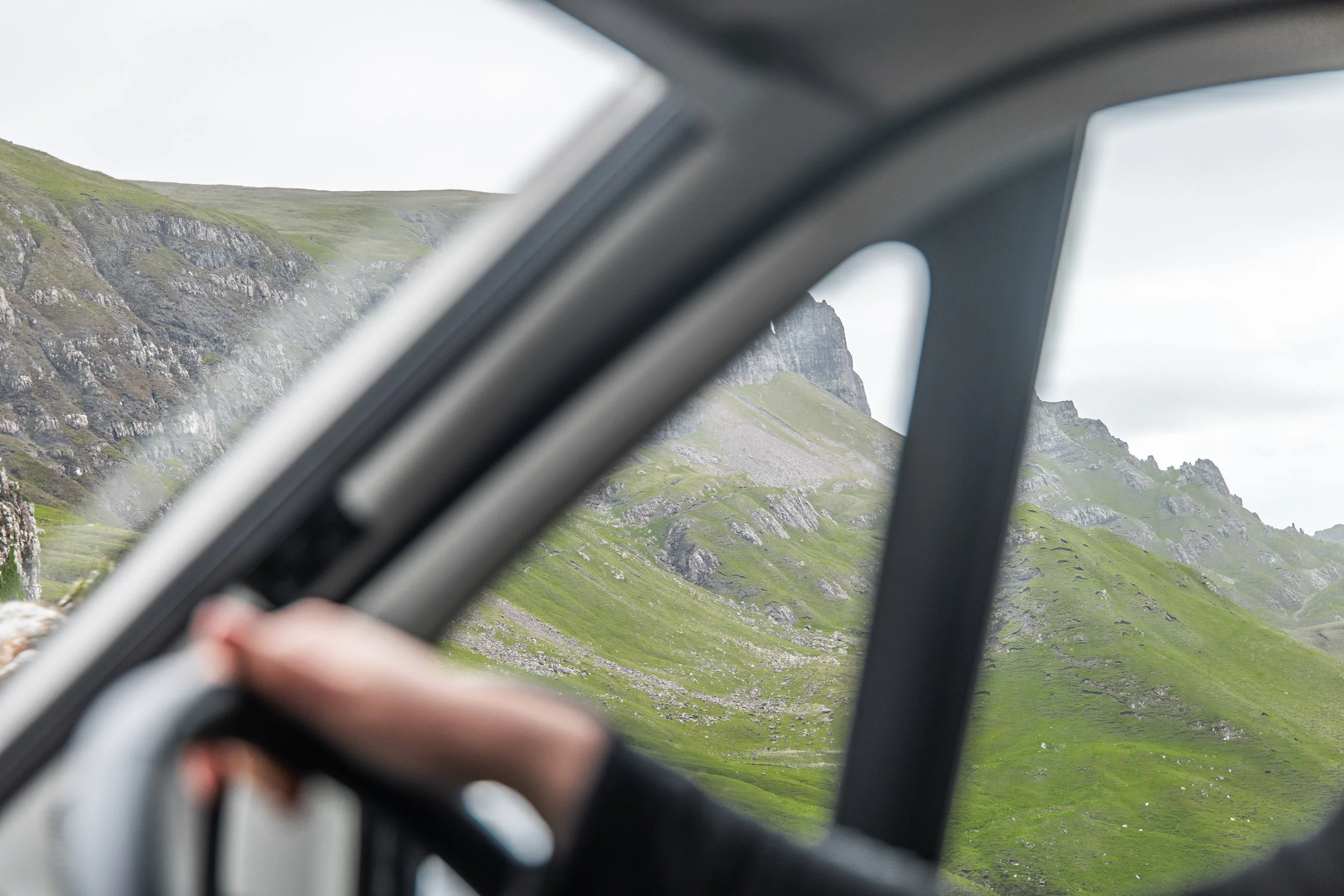A mountain landscape seen through a car window with a blurred hand in the foreground.