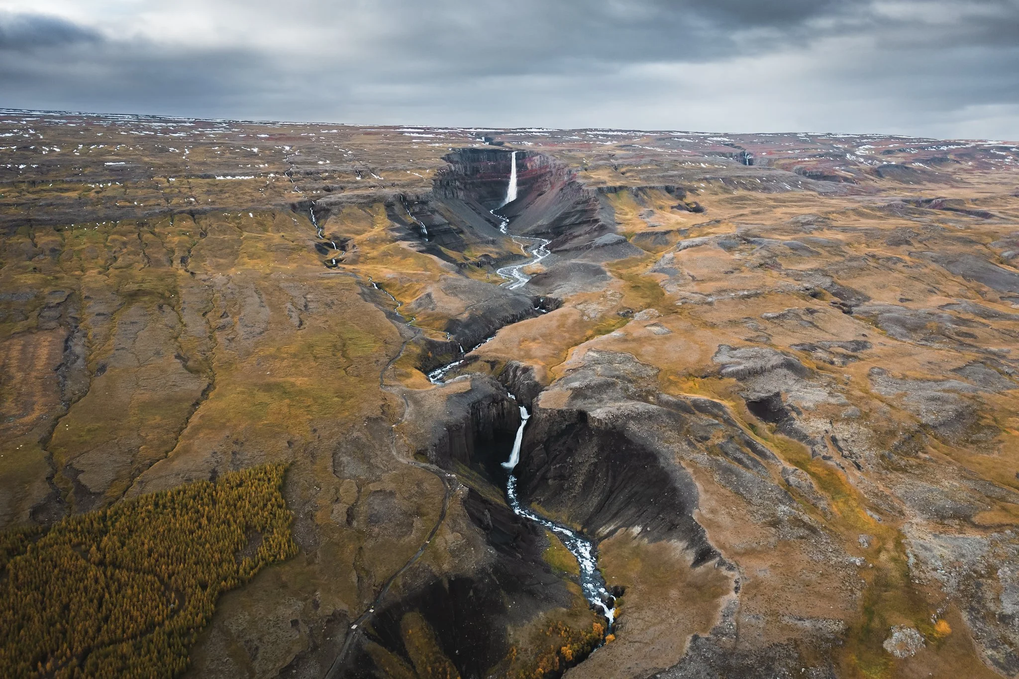 Aerial view of a canyon with multiple waterfalls, rocky terrain, sparse vegetation, and cloudy sky.