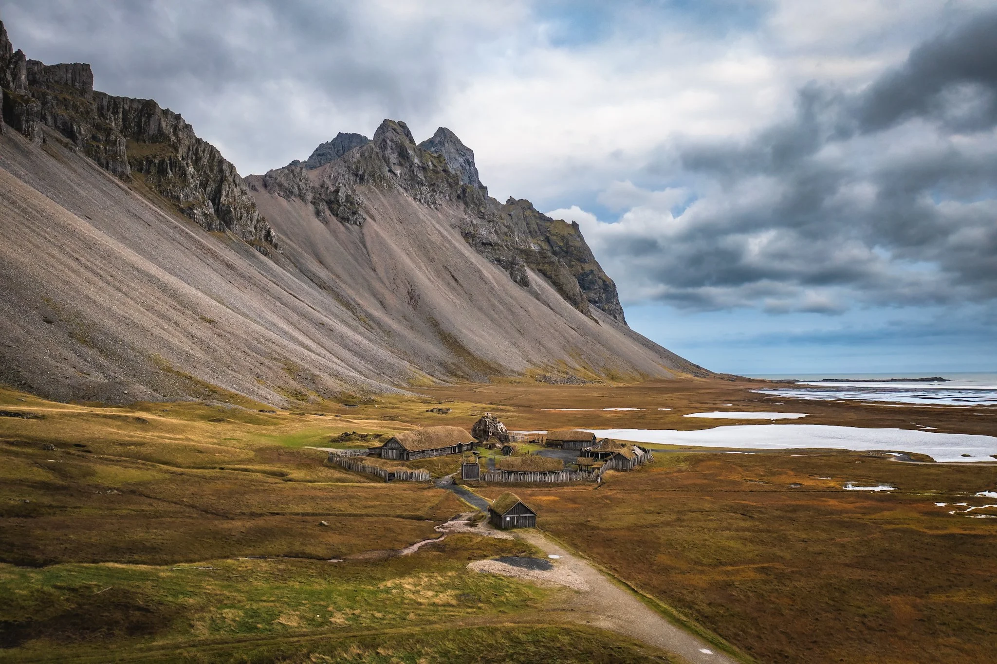 A landscape with a steep mountain slope on the left, a small cluster of wooden houses in the valley, and a body of water with marshy areas on the right, under a cloudy sky.
