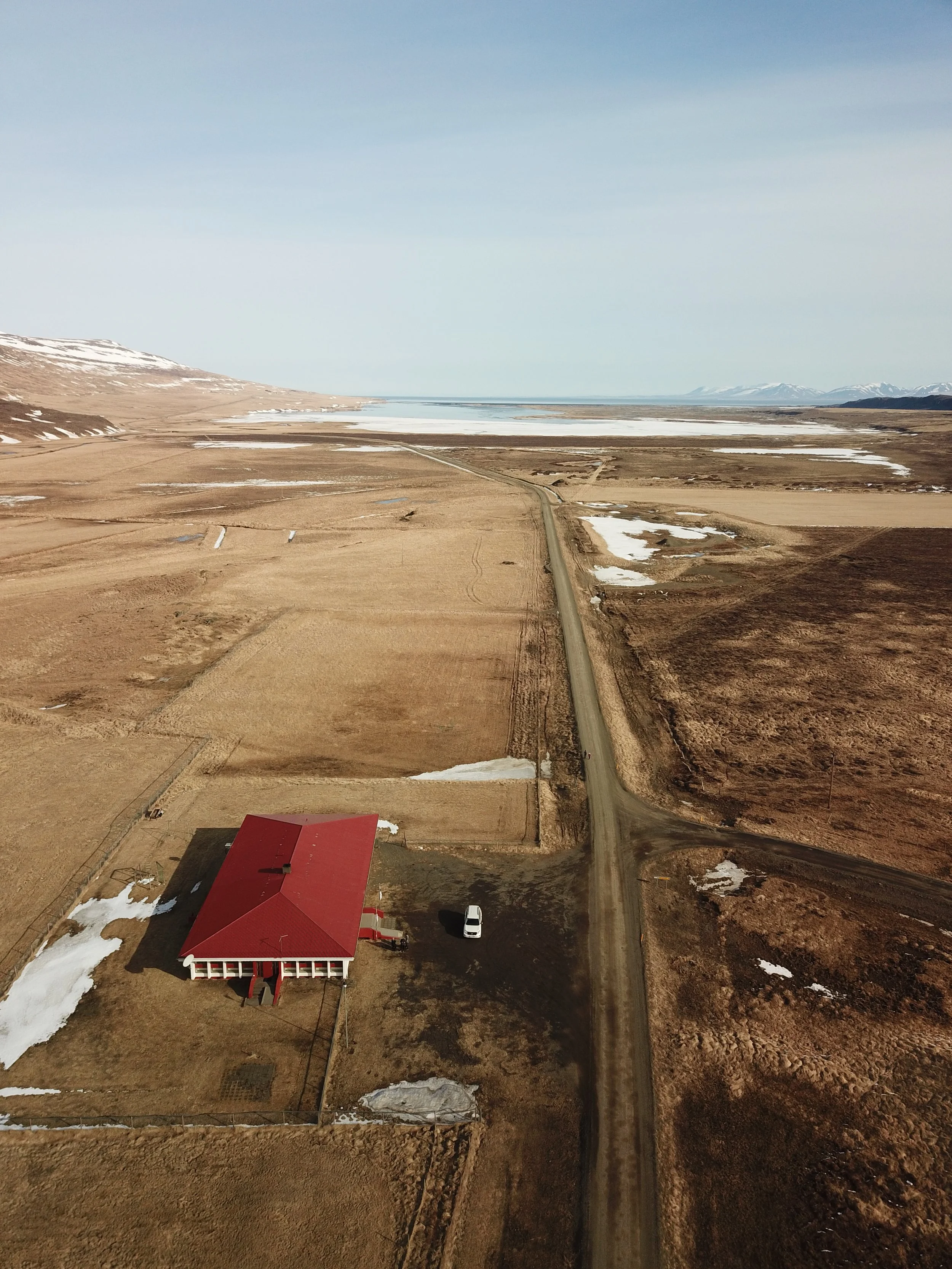 A rural landscape with a red-roofed house, a dirt driveway, open fields, patches of snow, a body of water in the distance, and mountains under a clear sky.