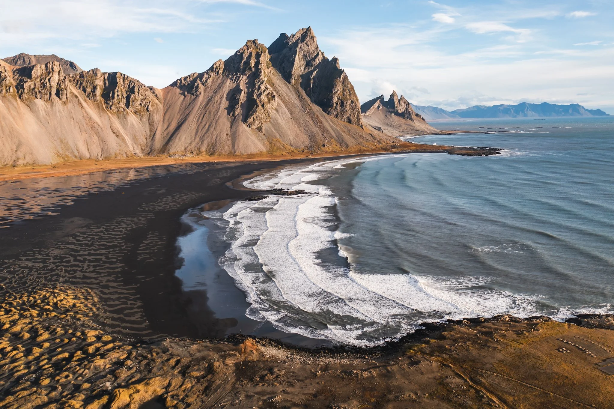 Scenic coastline with black sand beach, white waves, and rugged mountains in the distance under a partly cloudy sky.