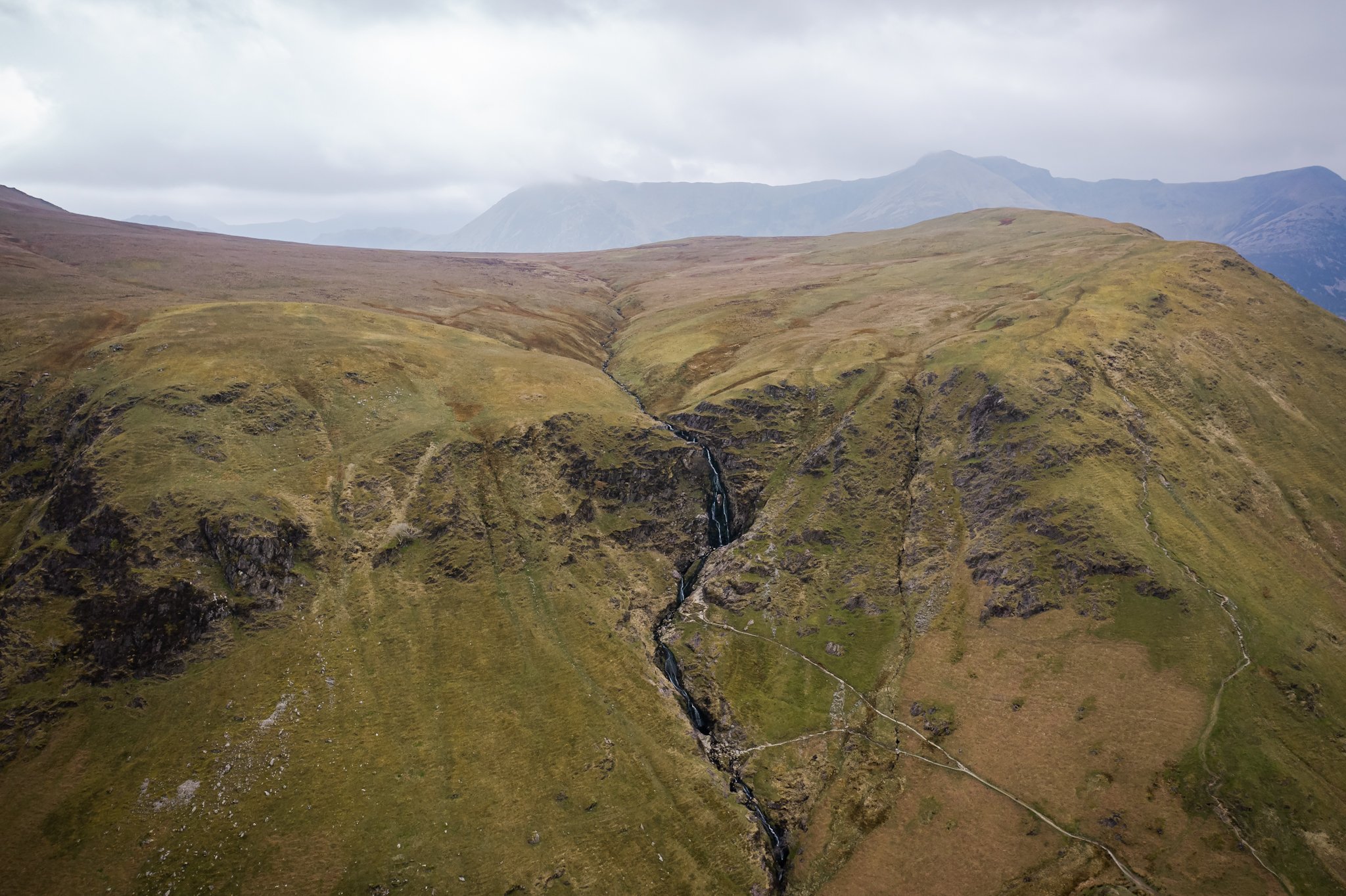Aerial view of green rolling hills with a small stream running through them, mountains in the background under a cloudy sky.