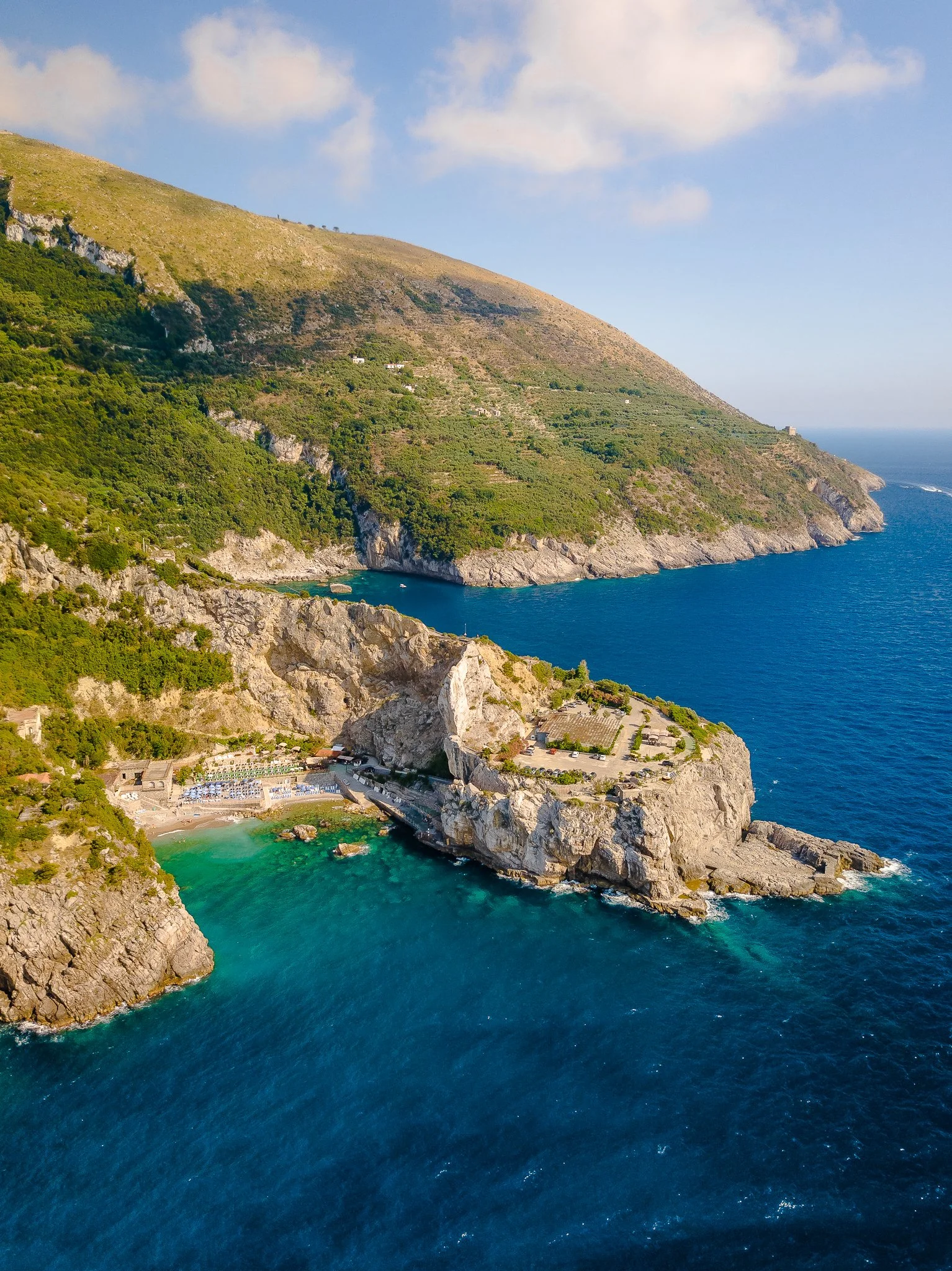 Aerial view of a scenic coastal area with green hills, rocky cliffs, a small beach with colorful umbrellas, and deep blue ocean water.