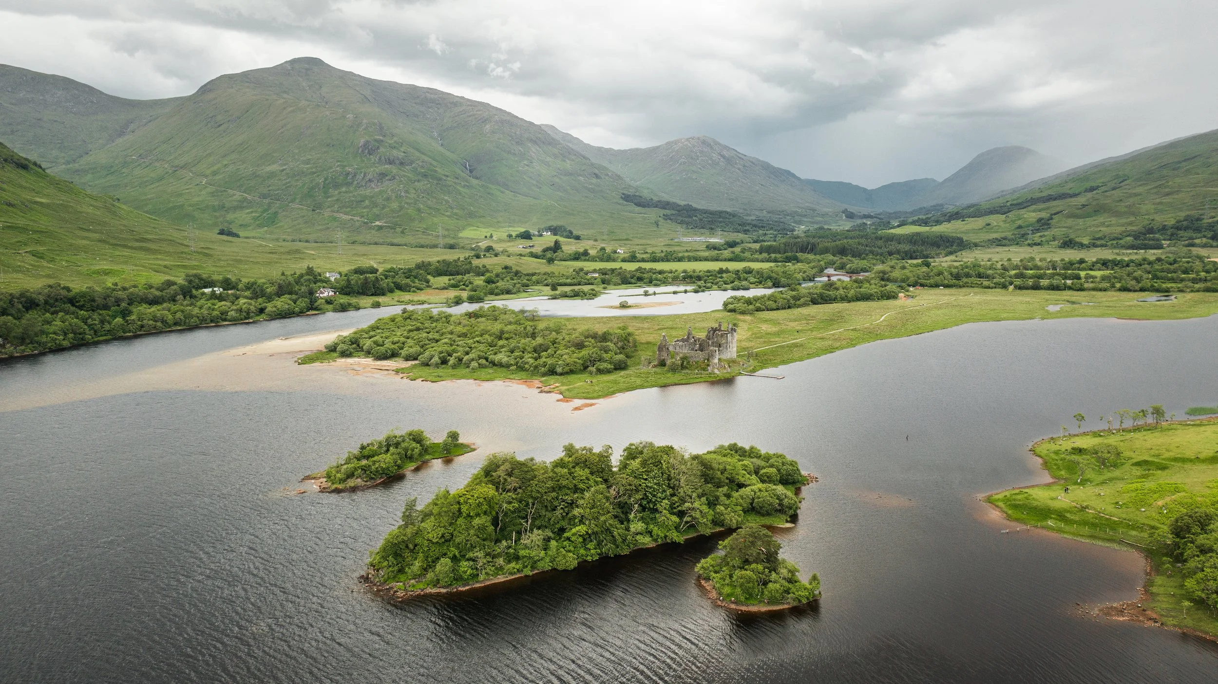 A scenic view of a lake with surrounding lush green trees, an old castle on a peninsula, green hills, and mountains in the background under a cloudy sky.