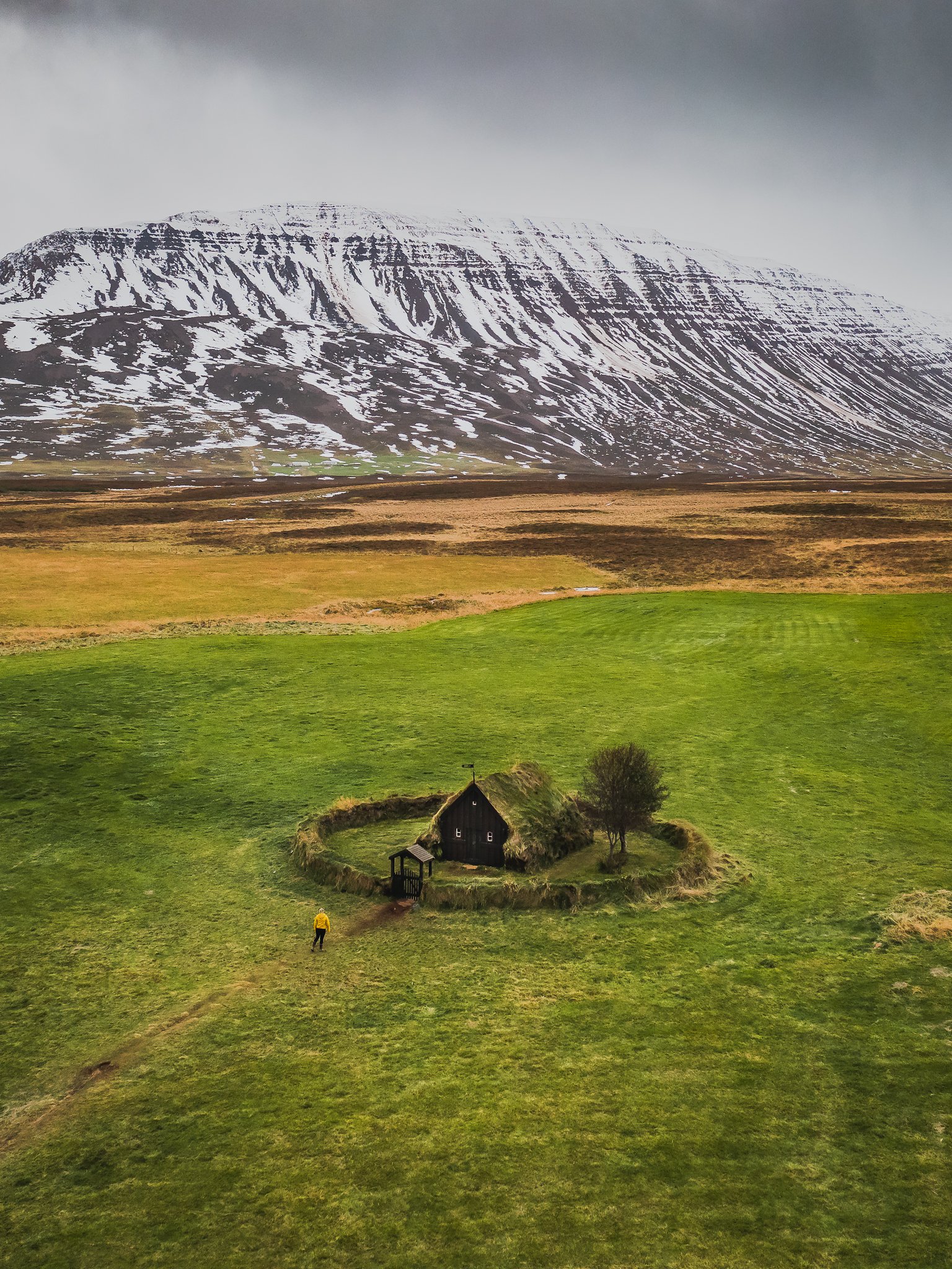 A small thatched-roof house surrounded by a circular hedge, with a person walking toward it across a green field, overlooking a large snow-capped mountain in the background.