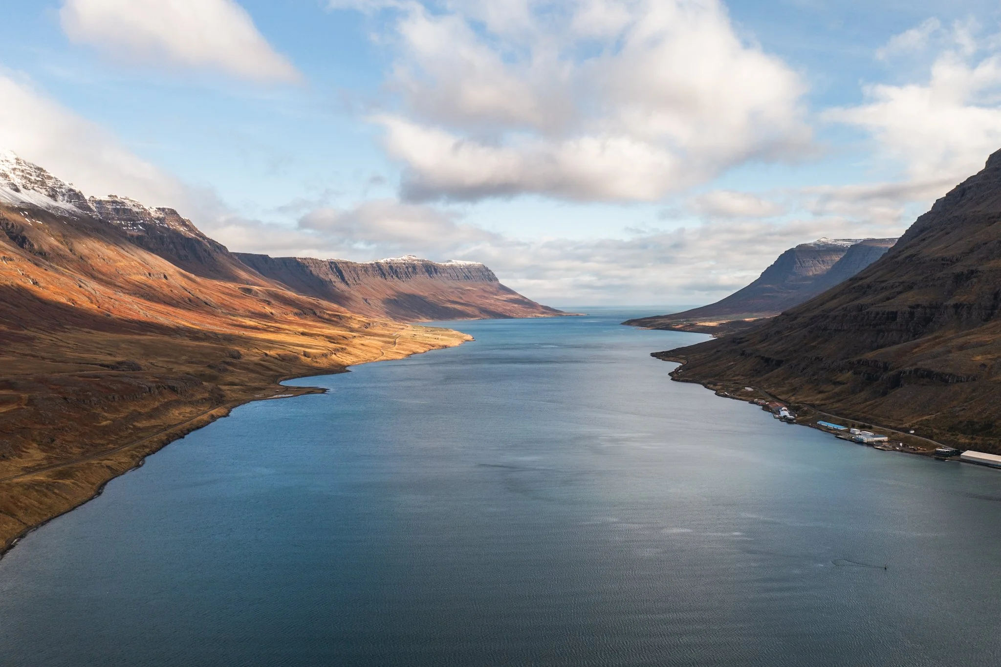 A scenic landscape featuring a wide river flowing between mountainous terrain, with snow-capped peaks under a partly cloudy sky.
