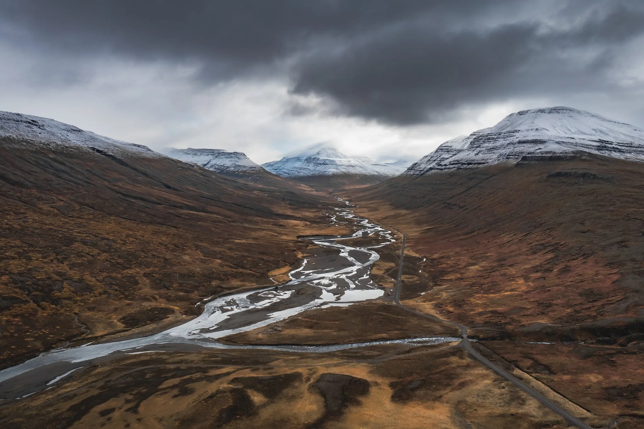 A landscape scene of a mountain valley with snow-capped mountains in the background. A winding river or stream runs through the brown and green valley floor. The sky is overcast with dark clouds, creating a moody atmosphere.