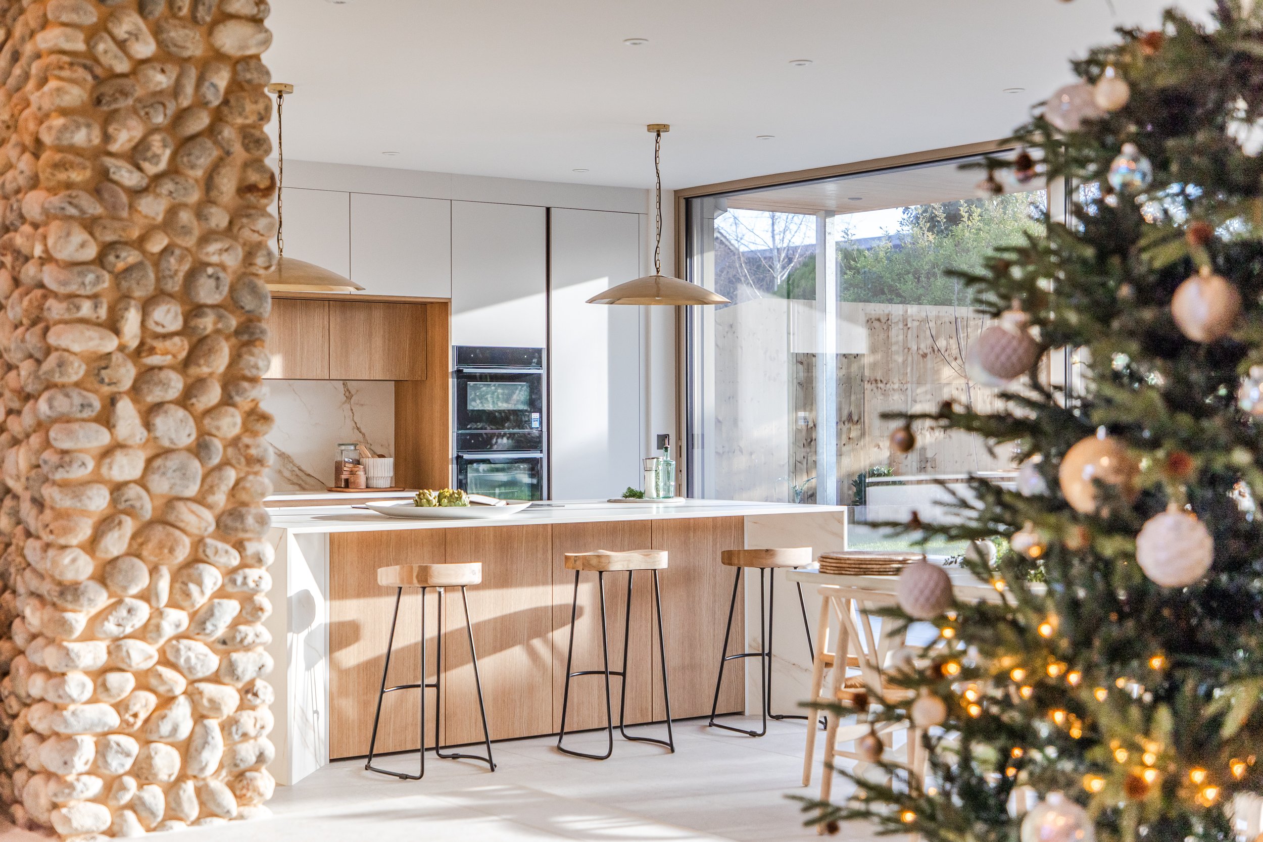 Modern kitchen with white and wood cabinets, pendant lights, a large window, and a decorated Christmas tree.