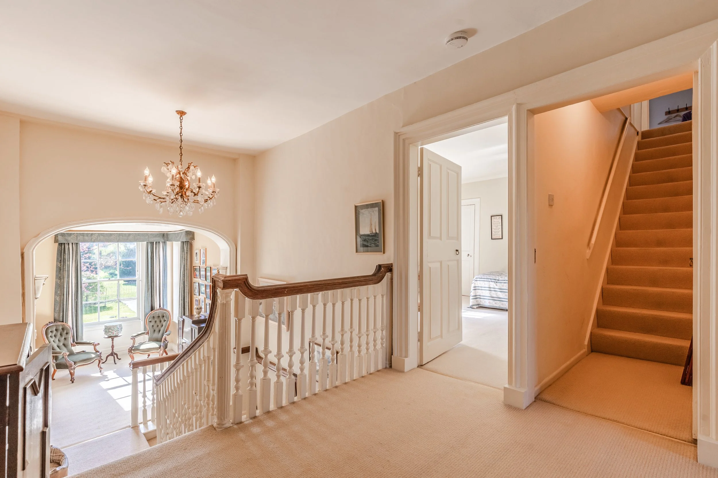 An interior view of a house showing staircase, hallway, doorway, chandelier, and a sitting area with chairs and a window.