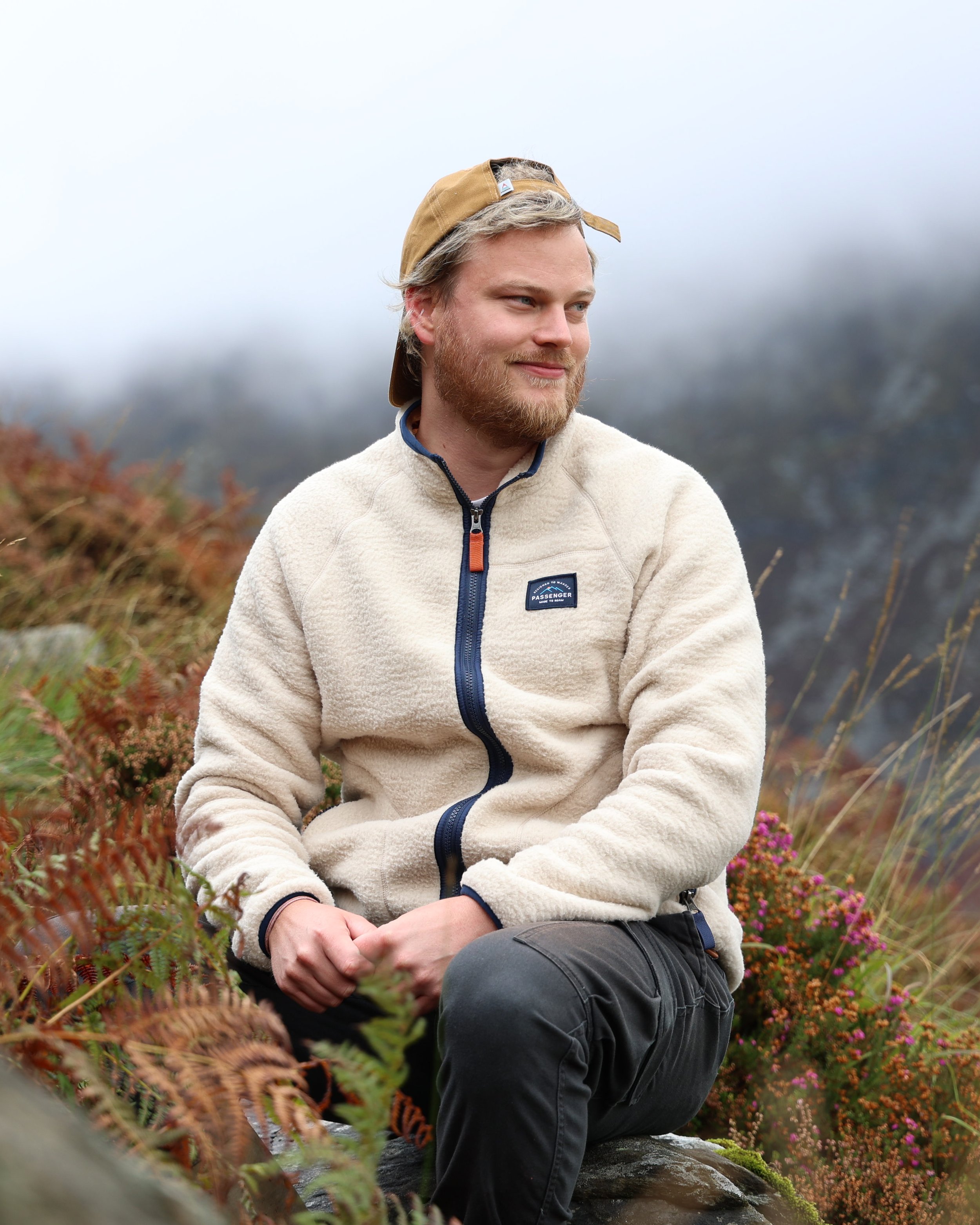 A man with blonde hair and a beard, wearing a tan cap backwards, a cream fleece jacket, and dark pants, is sitting outdoors on a rock amidst a natural setting with bushes and trees, under a cloudy sky.