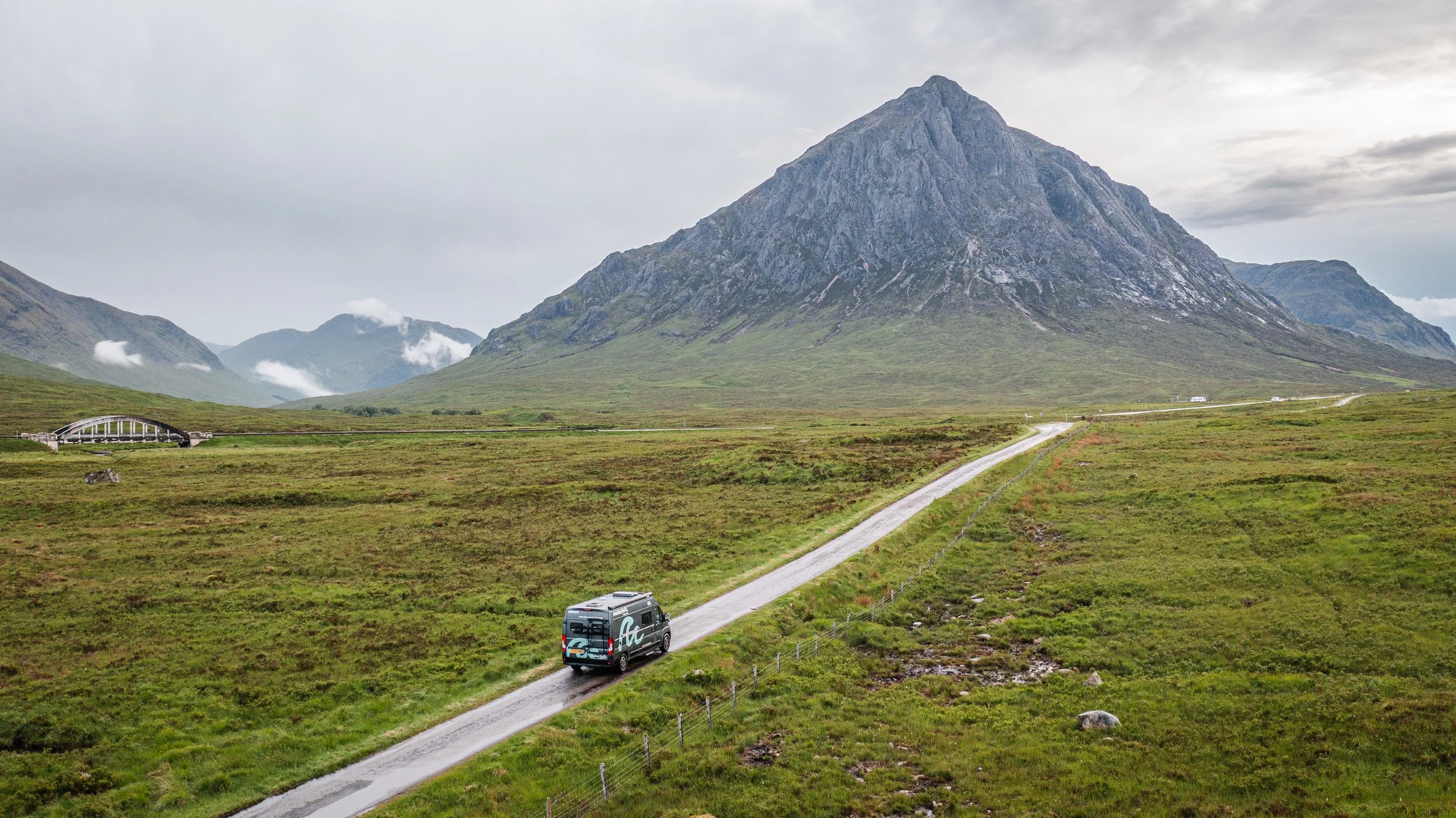 A lone vehicle driving along a winding dirt road through a green, open landscape with a tall mountain in the background, under a cloudy sky.