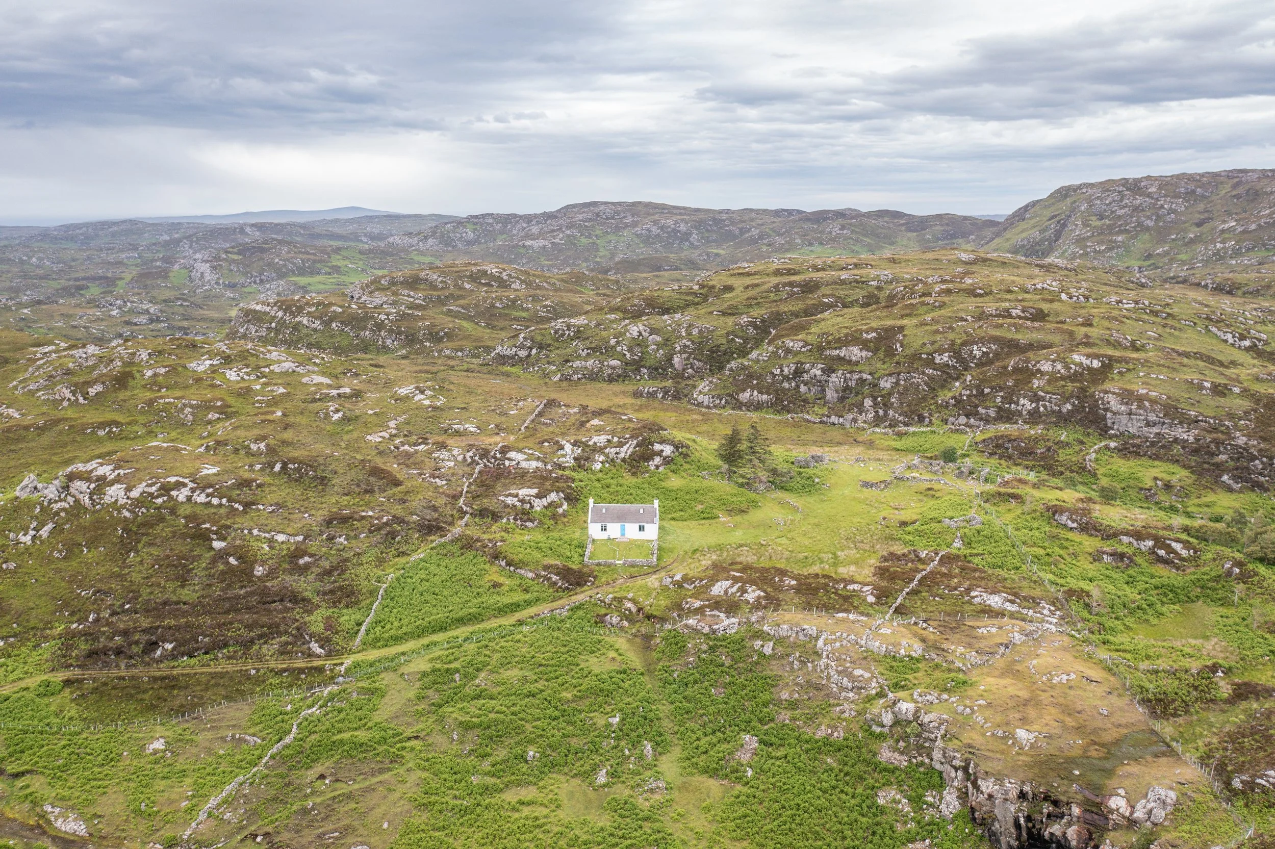 A small white house with a blue door situated in a rural, hilly landscape with rocky terrain and patches of green vegetation under a cloudy sky.