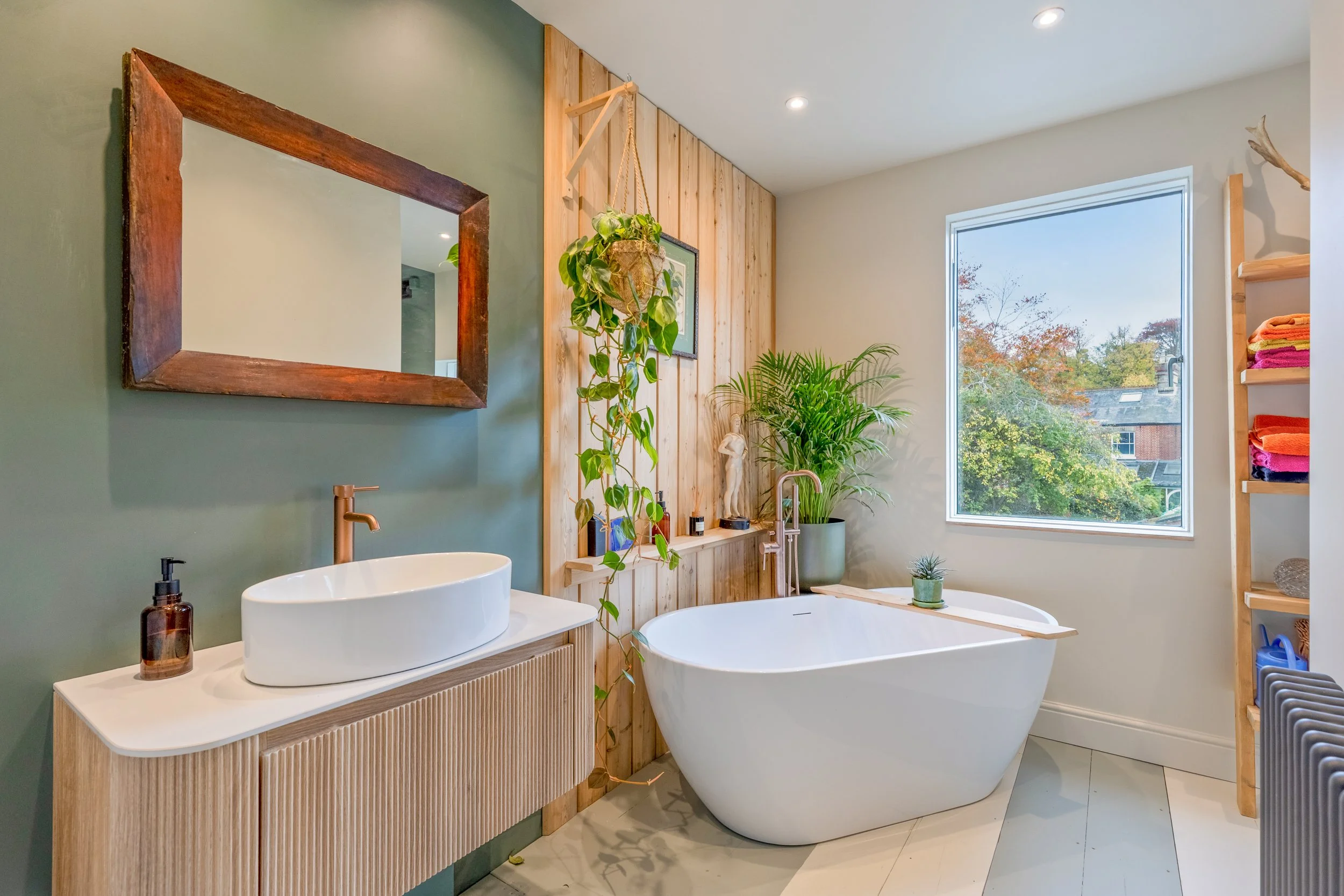 Modern bathroom featuring a large white bathtub, a wooden vanity with vessel sink, green accent wall, indoor plants, a square mirror with a wooden frame, and a window showing trees outside.
