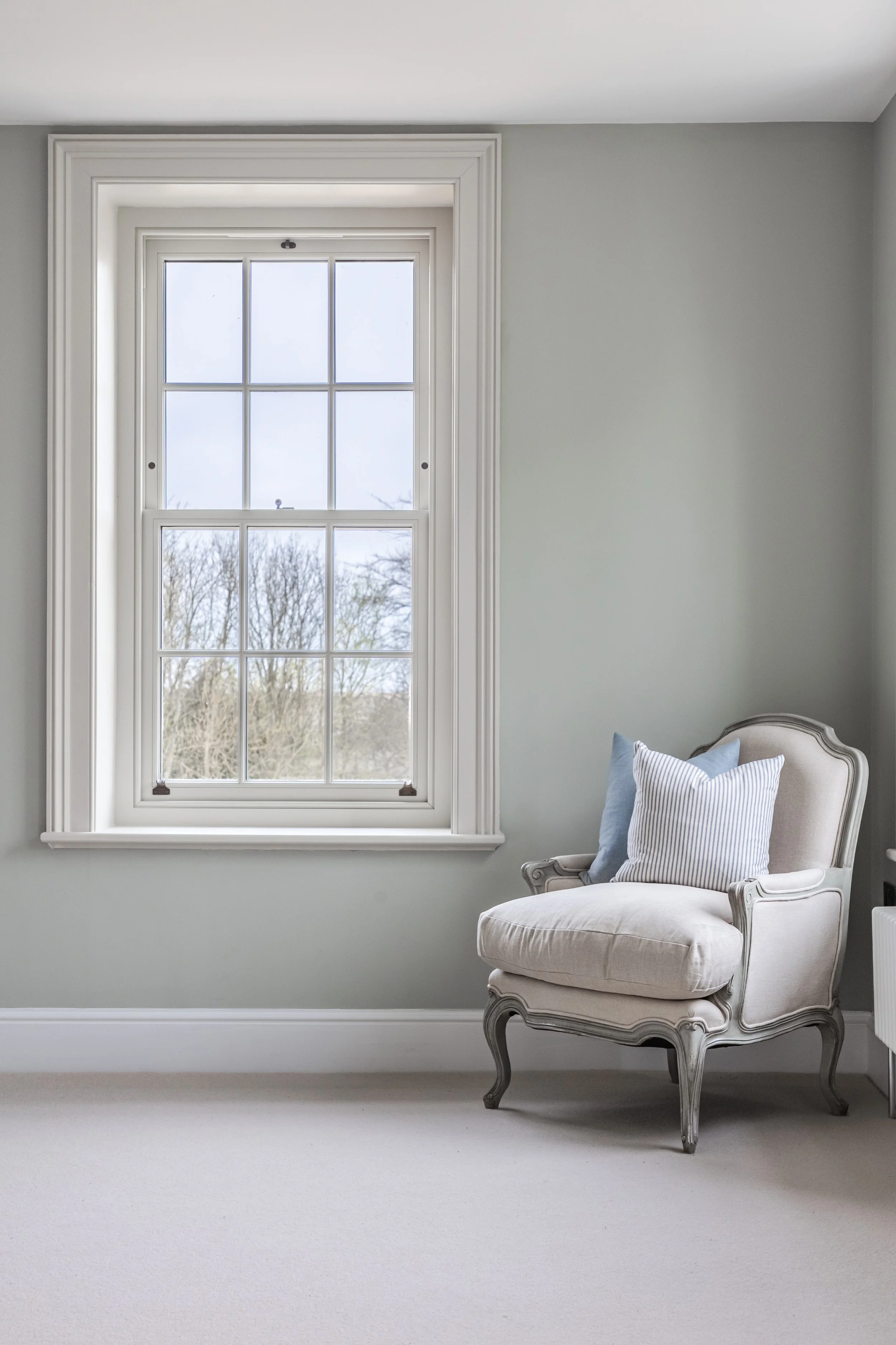 A white vintage armchair with light blue and striped pillows, positioned next to a large window with white trim, in a light gray room.