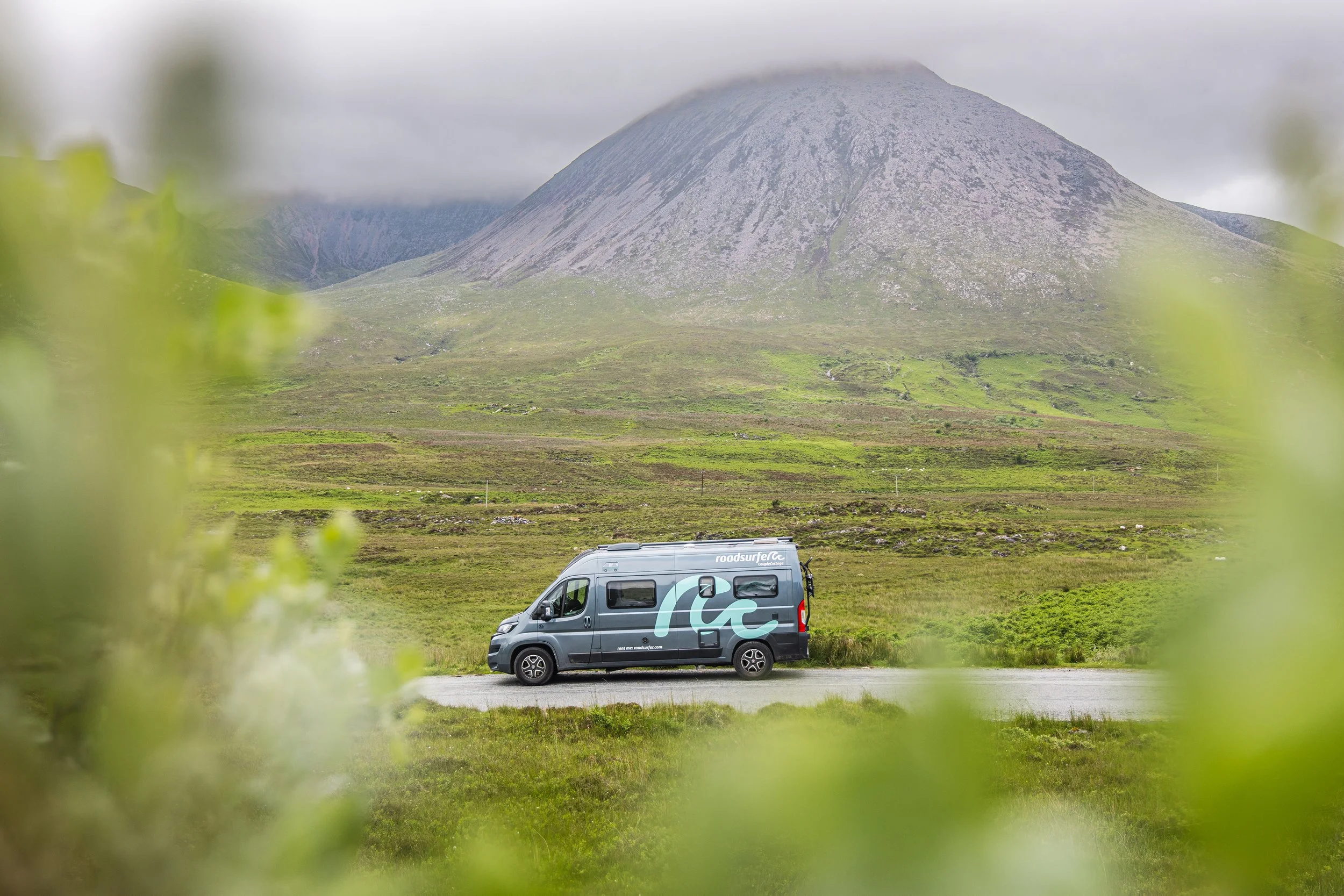 A gray camper van parked on green grass in front of a mountain with a cloud-covered peak