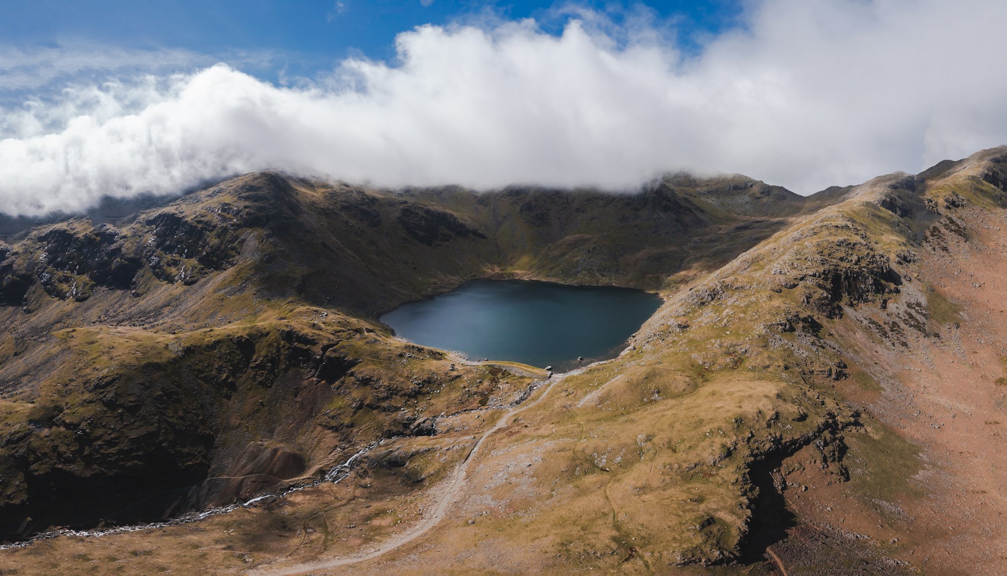 Mountain landscape with a small lake and flowing streams, partly covered by clouds under a partly cloudy blue sky.