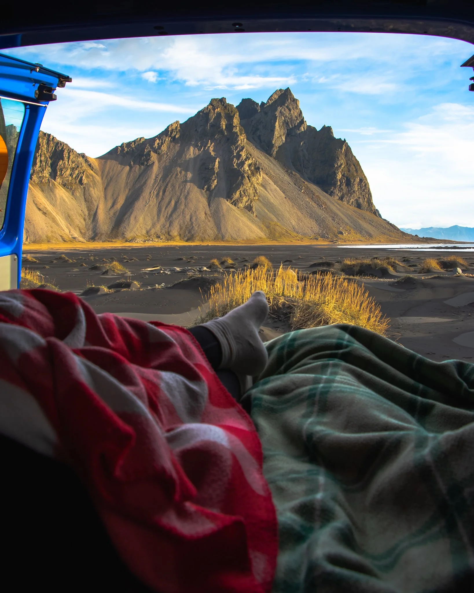 Person lying inside a vehicle with a mountain landscape and partly cloudy sky visible through the window, with legs and feet extended outward.