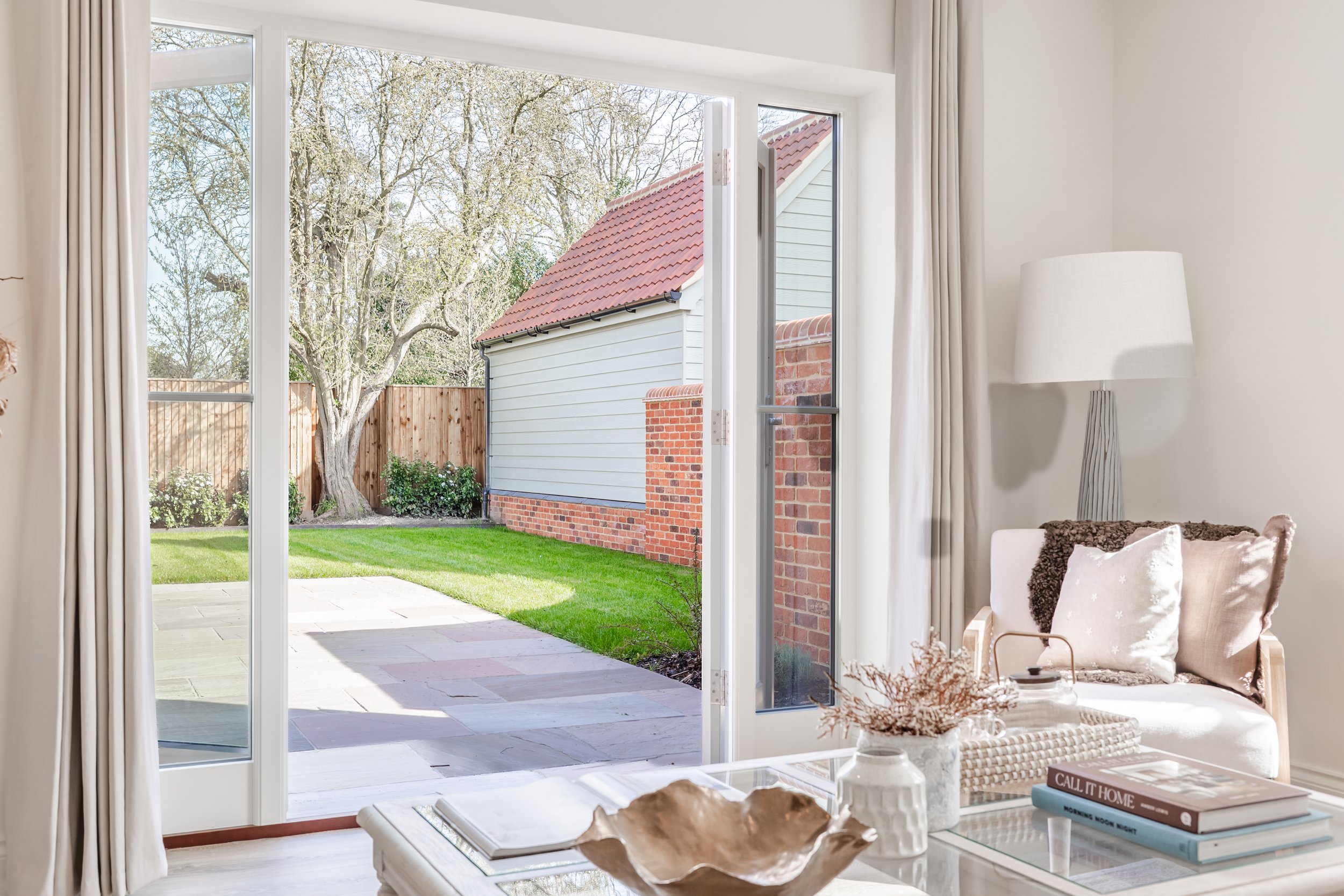 Interior view of a living room looking out through large open glass sliding doors onto a backyard with a lawn, patio, wooden fence, a tree, and neighboring house with a red-tiled roof.