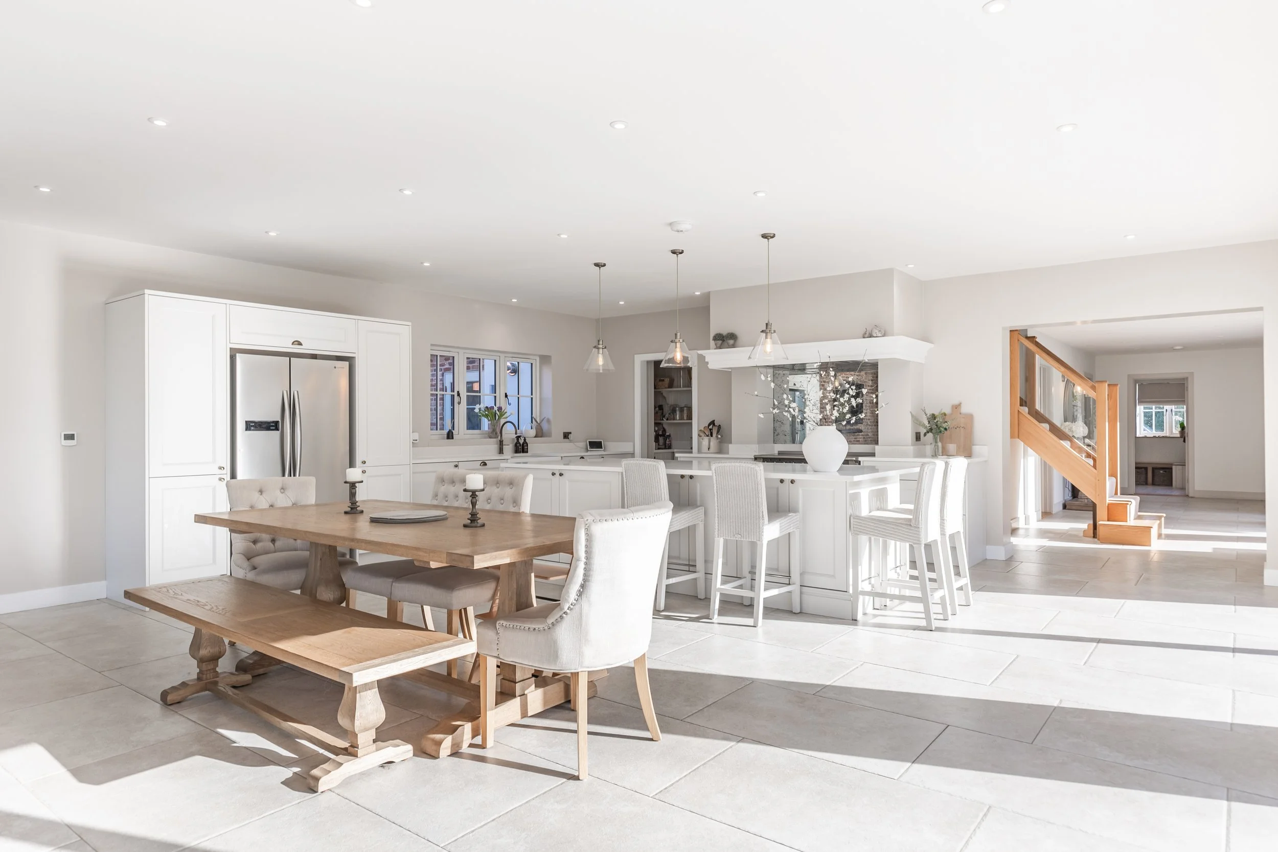 Bright, modern open-concept kitchen and dining area with white cabinetry, a large wooden dining table, white upholstered chairs, and a breakfast bar with white stools. The space features pendant lighting, a stainless steel refrigerator, large windows