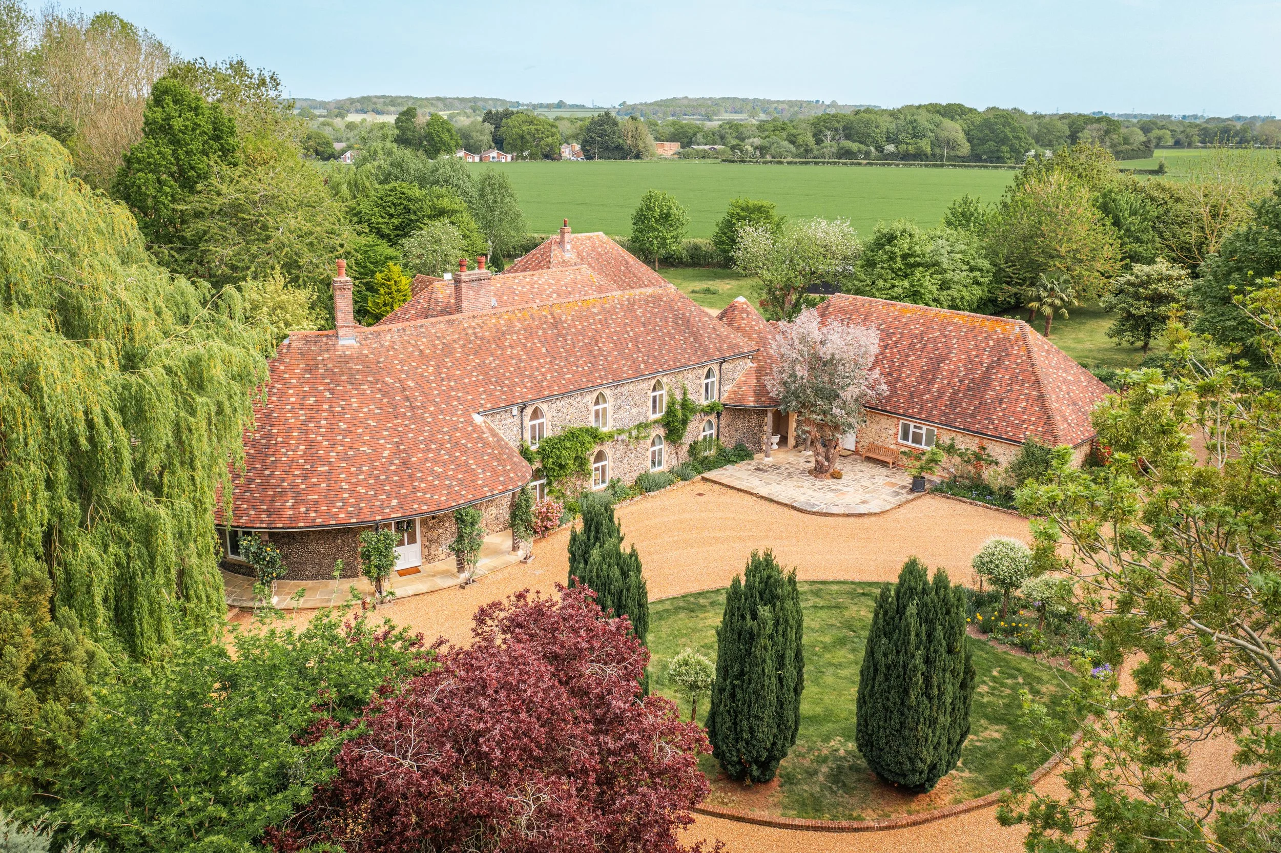 Aerial view of a large house with a red tile roof surrounded by lush green trees and garden, overlooking fields and countryside.