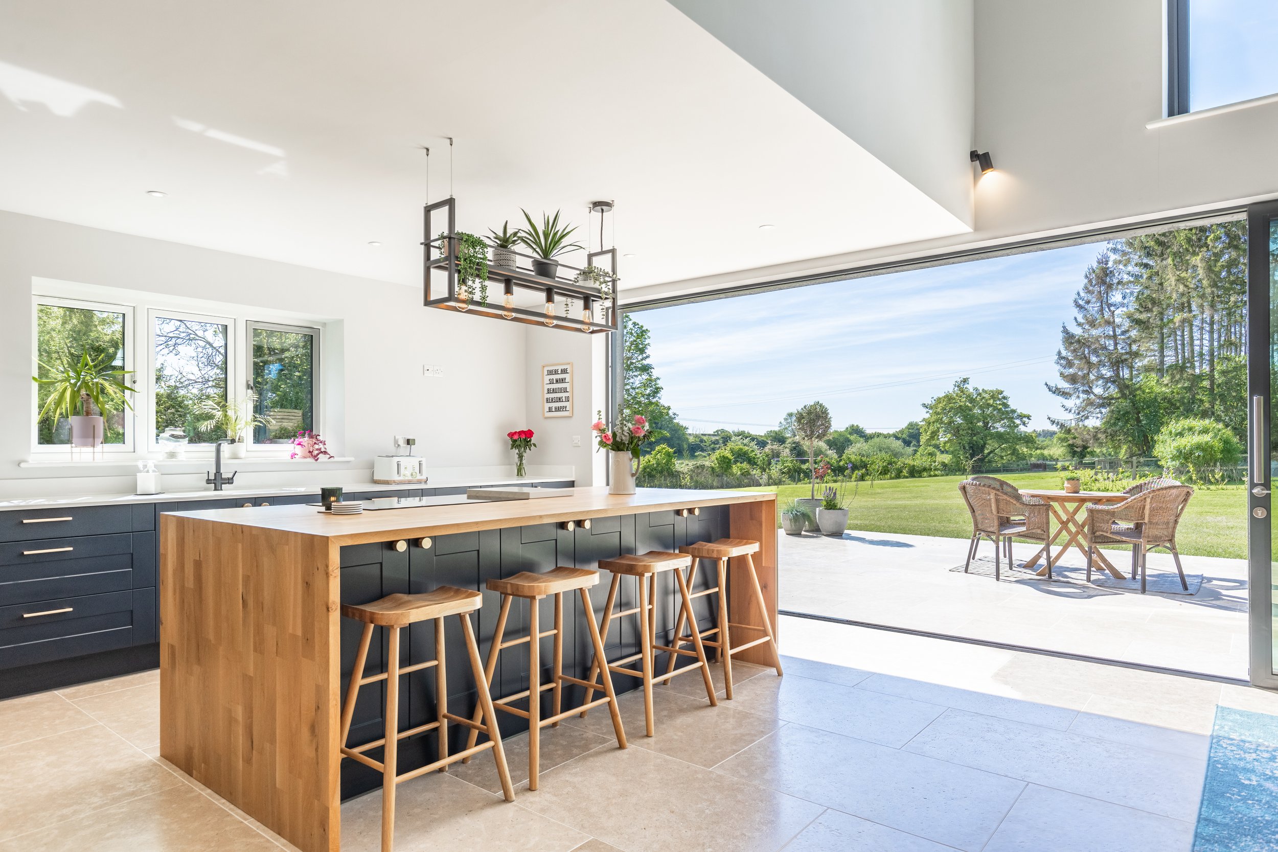 A modern kitchen with a large window and sliding glass door opening to a grassy outdoor area. The kitchen features a wooden island with navy blue cabinets, four wooden barstools, and potted plants on the counters and shelves. Outside, there is a roun