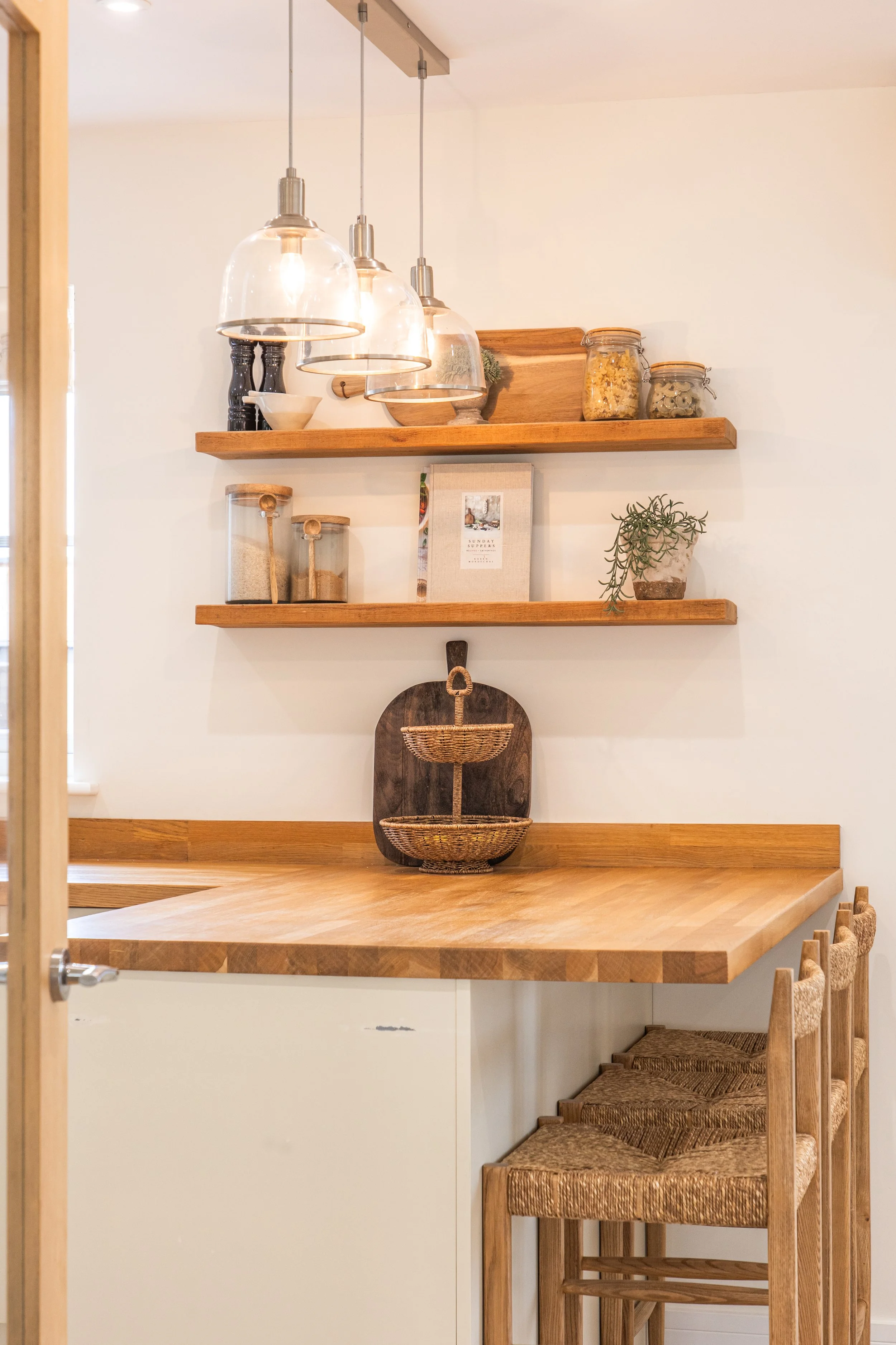 A wooden kitchen countertop with three barstools with woven seats beneath it. Two wooden shelves above hold various jars, bowls, and plants, with a hanging light fixture with three glass shades overhead.