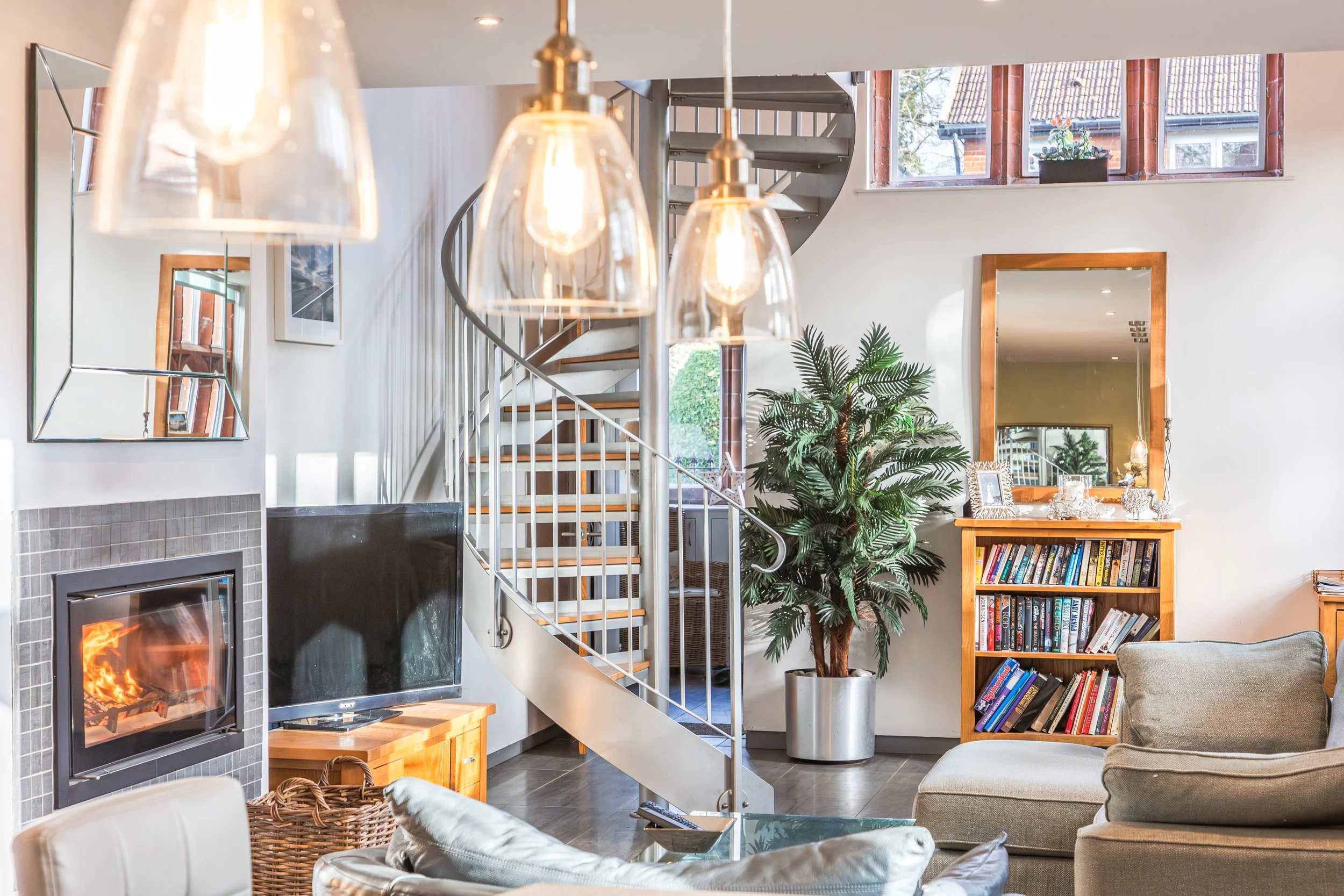 Living room with a fireplace, a television, a spiral staircase, a large mirror, a bookshelf filled with books, a large potted plant, and windows with a view of houses outside.