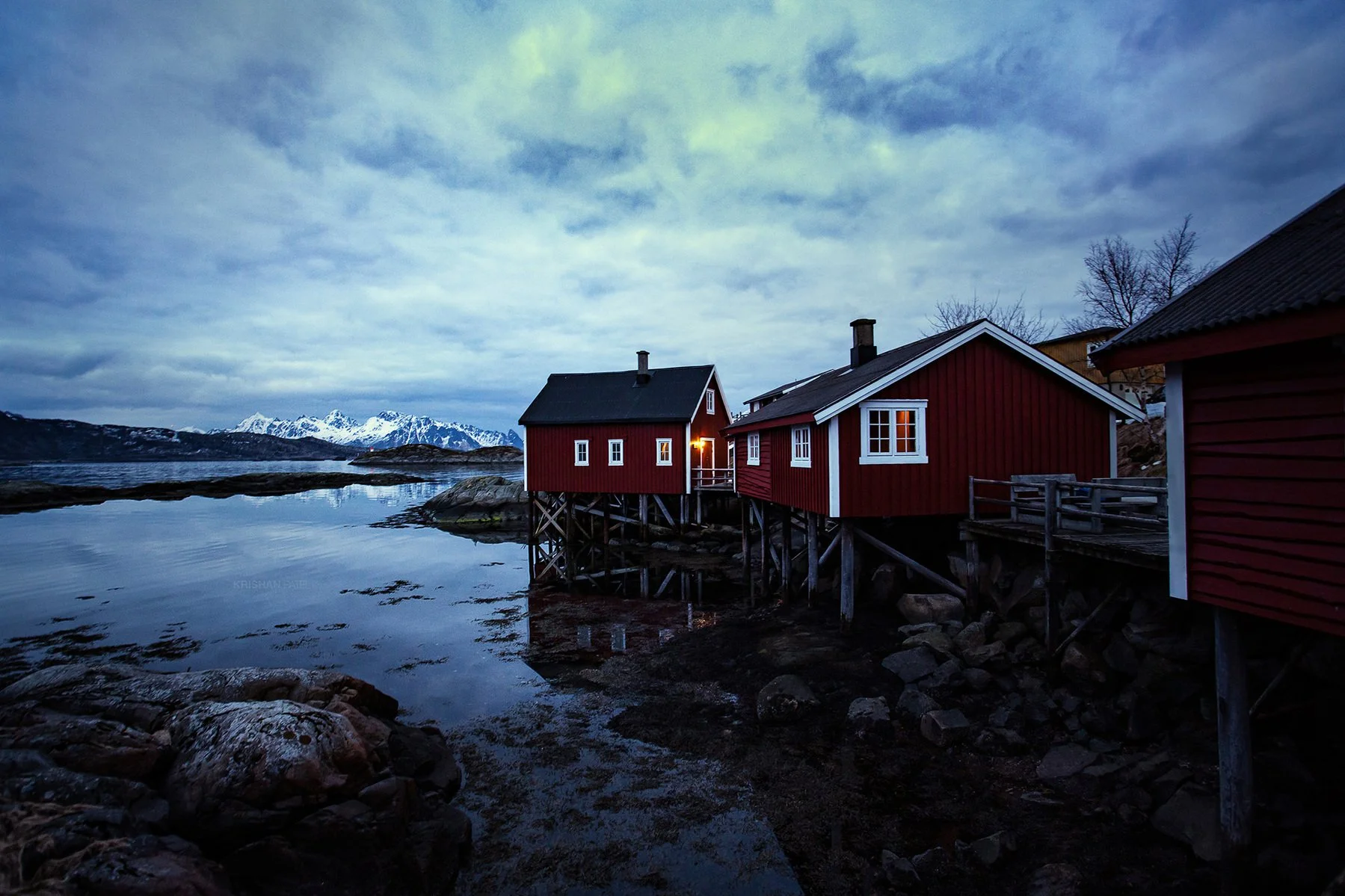Red wooden houses built on stilts over water near rocky shoreline with snow-capped mountains in distance under cloudy sky during dusk or dawn.