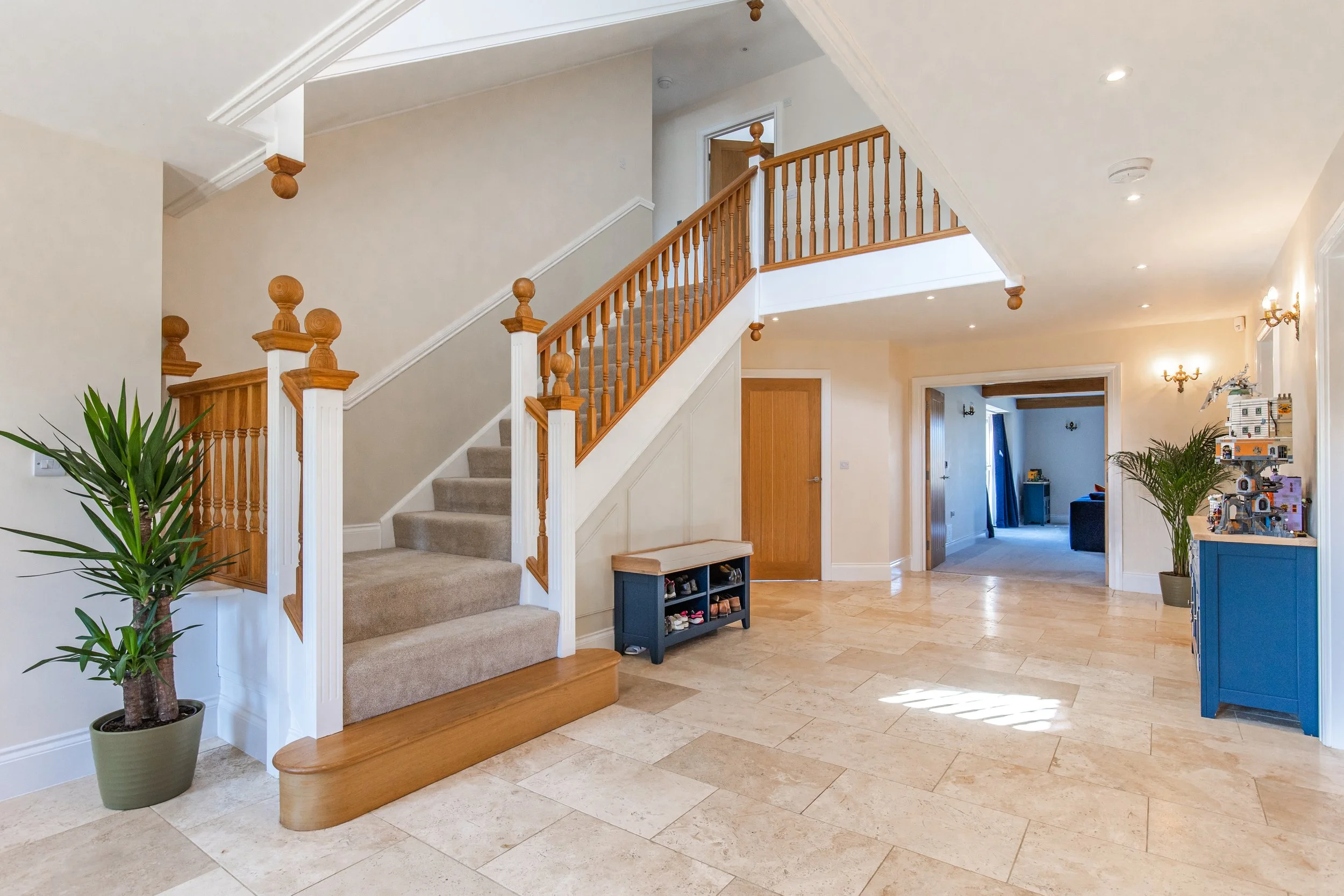 Interior view of a residential home with a staircase, wooden banister, beige walls, tiled flooring, and some indoor plants. There is a doorway leading to another room with visible play area and furniture.