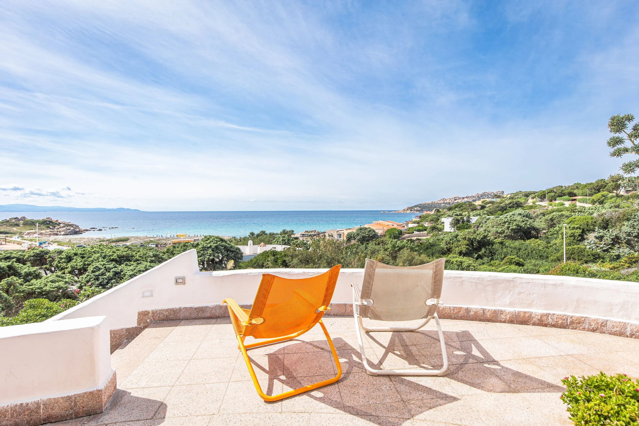 A scenic view of the ocean and coastline from a balcony with colorful chairs, green trees, and a clear blue sky with wispy clouds.