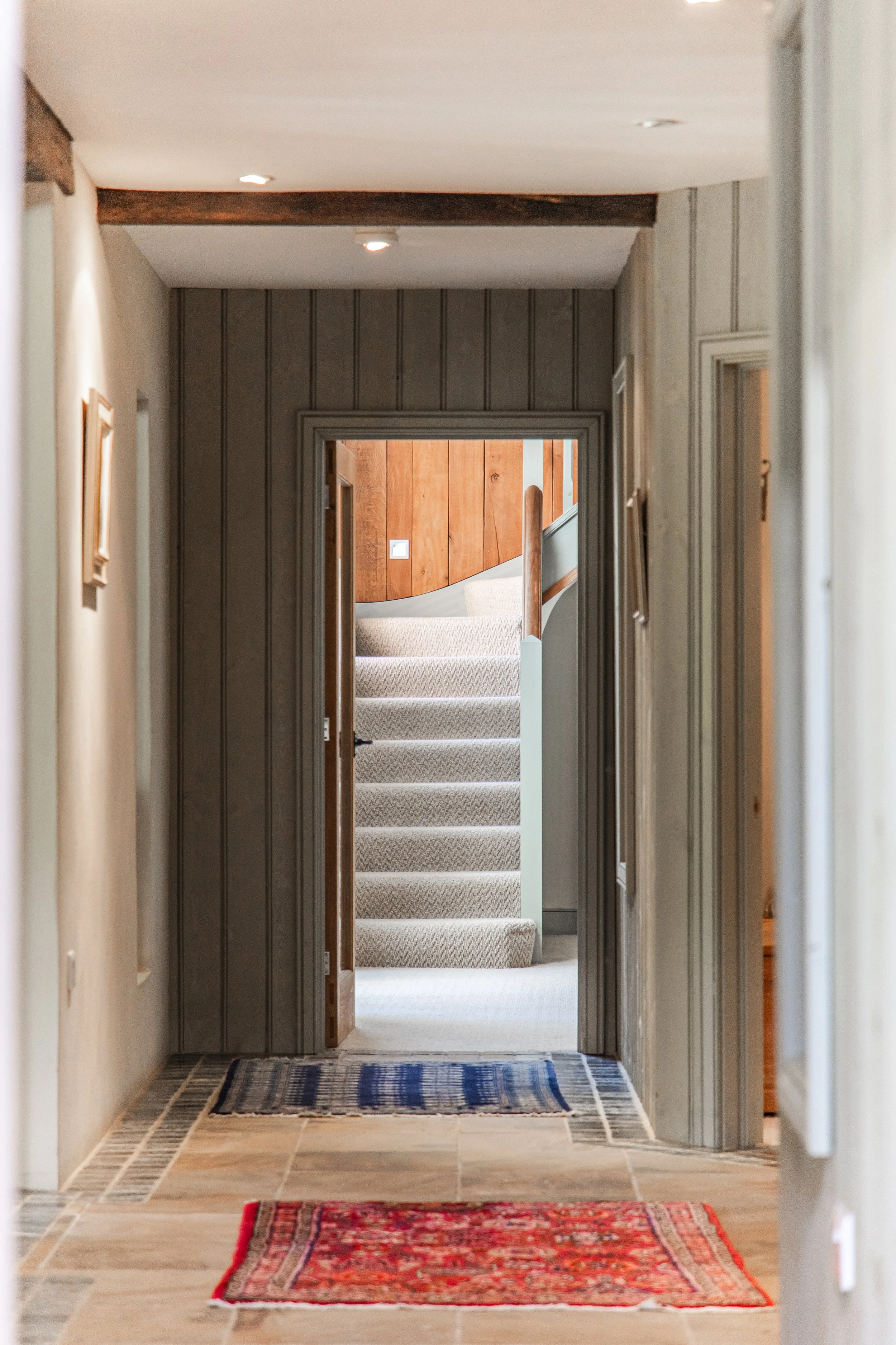 A hallway with woven rugs, leading to a staircase with beige carpet and wooden paneling background.