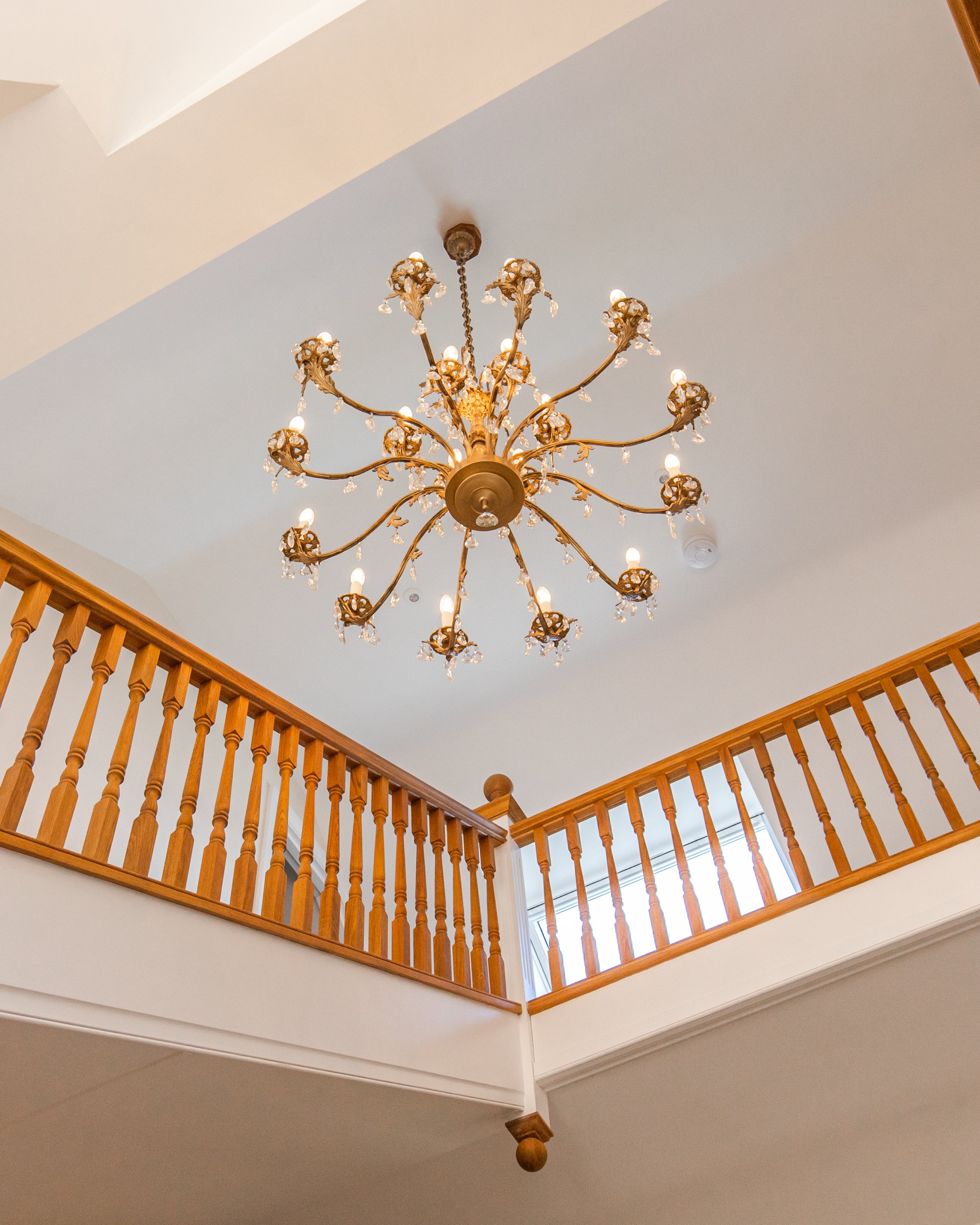 Interior view of a two-story house showing a chandelier hanging from the ceiling, with wooden railing on the upper level, and a window in the background.