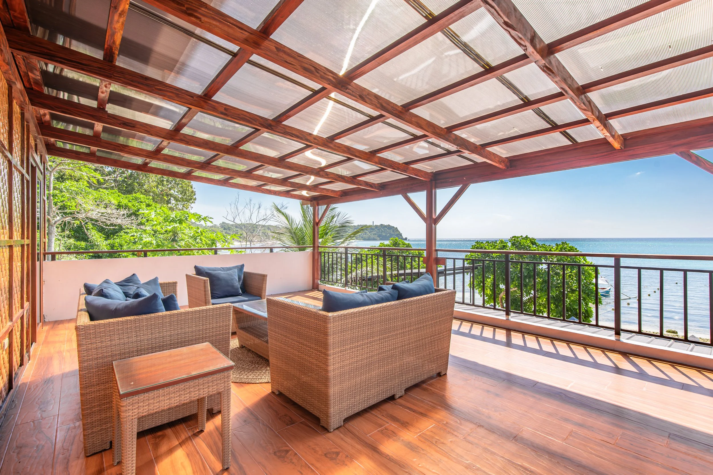Outdoor balcony with wicker furniture and blue cushions overlooking the ocean with green trees and a distant hill.