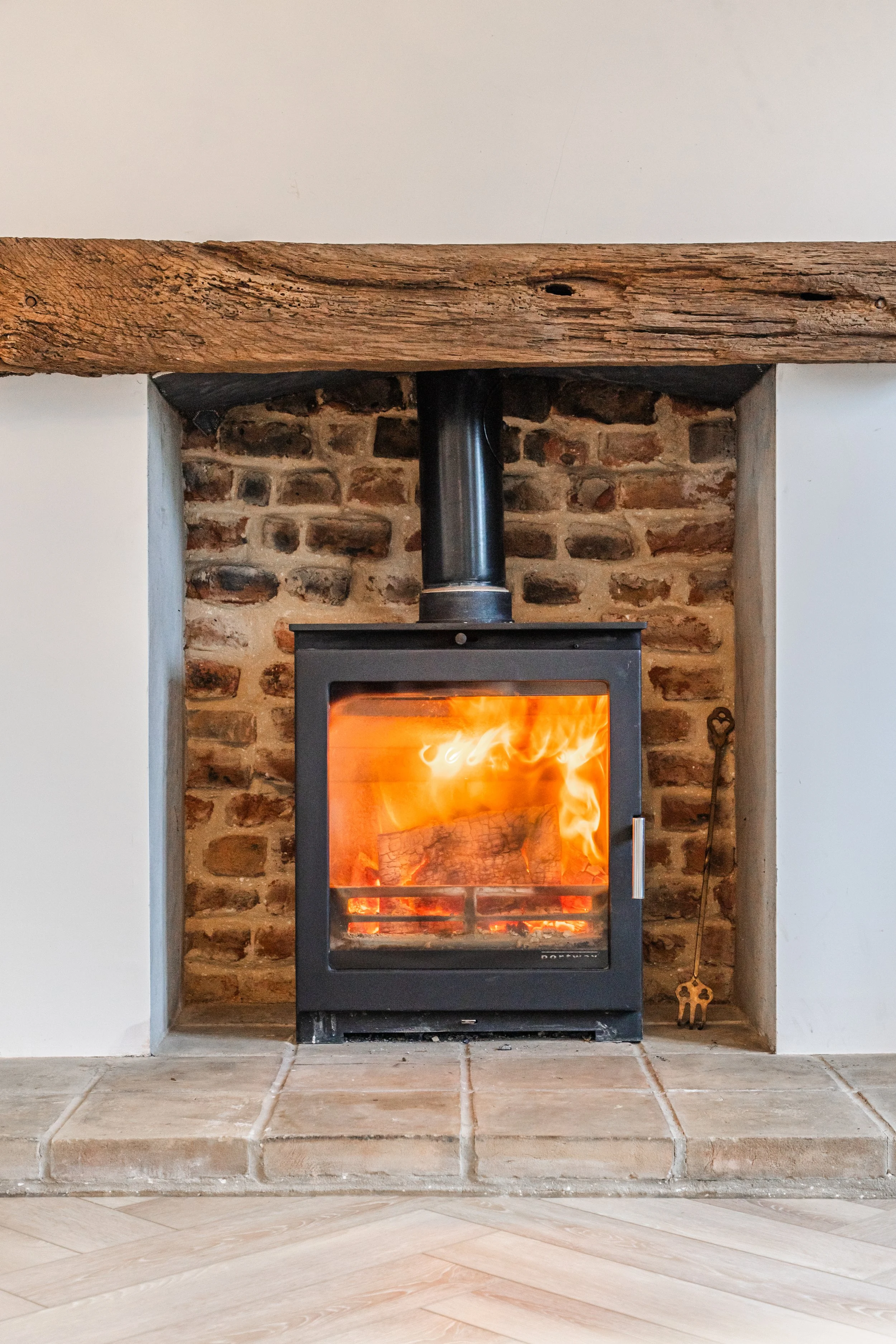 A fireplace with a wood stove burning inside, set against a brick wall. The fireplace is framed by a wooden beam on top and a concrete surround. The floor in front is tiled.