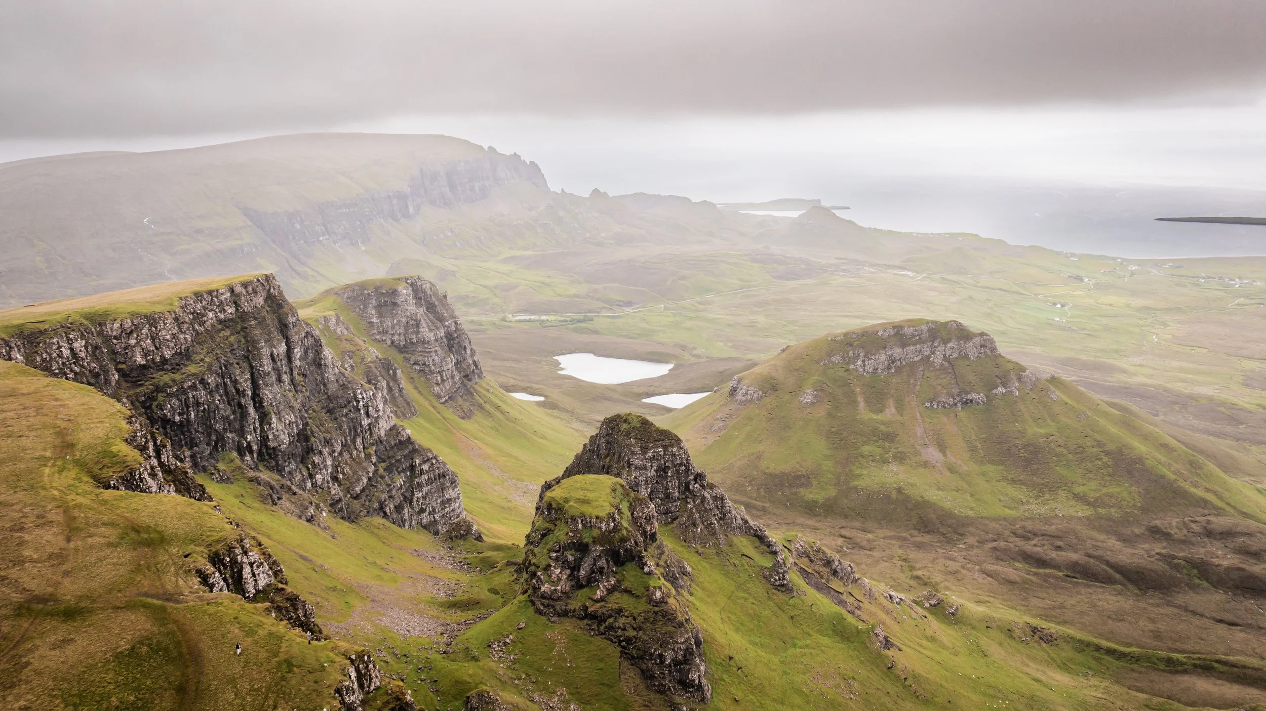 A scenic view of green rolling hills and rugged cliffs with small lakes, under a cloudy sky in a coastal landscape.