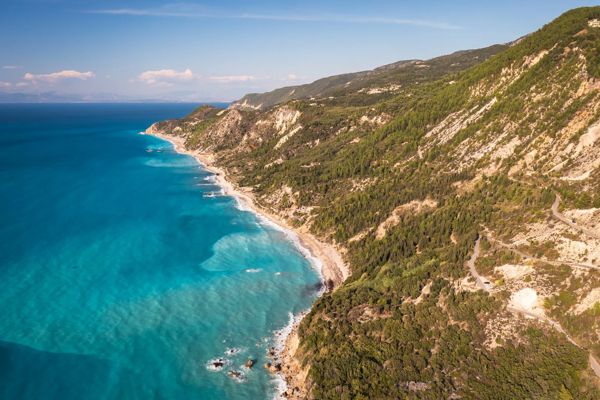 Aerial view of a coastline with turquoise water, sandy beach, green forested cliffs, and winding roads along the hillside.