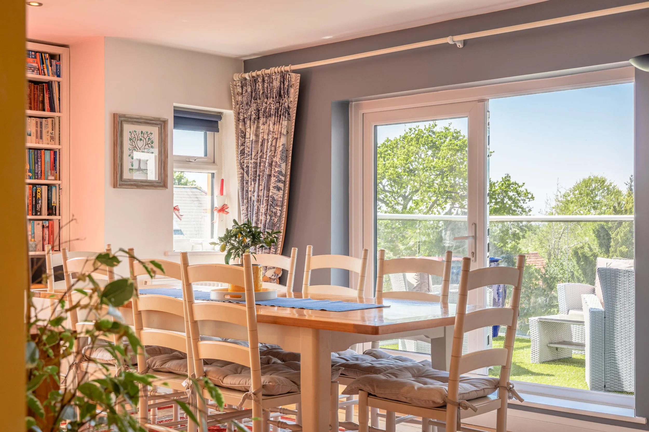 Bright dining room with large windows showing green trees outside, wooden table surrounded by chairs with cushions, bookshelf filled with books, wall art, and curtains with a blue pattern.