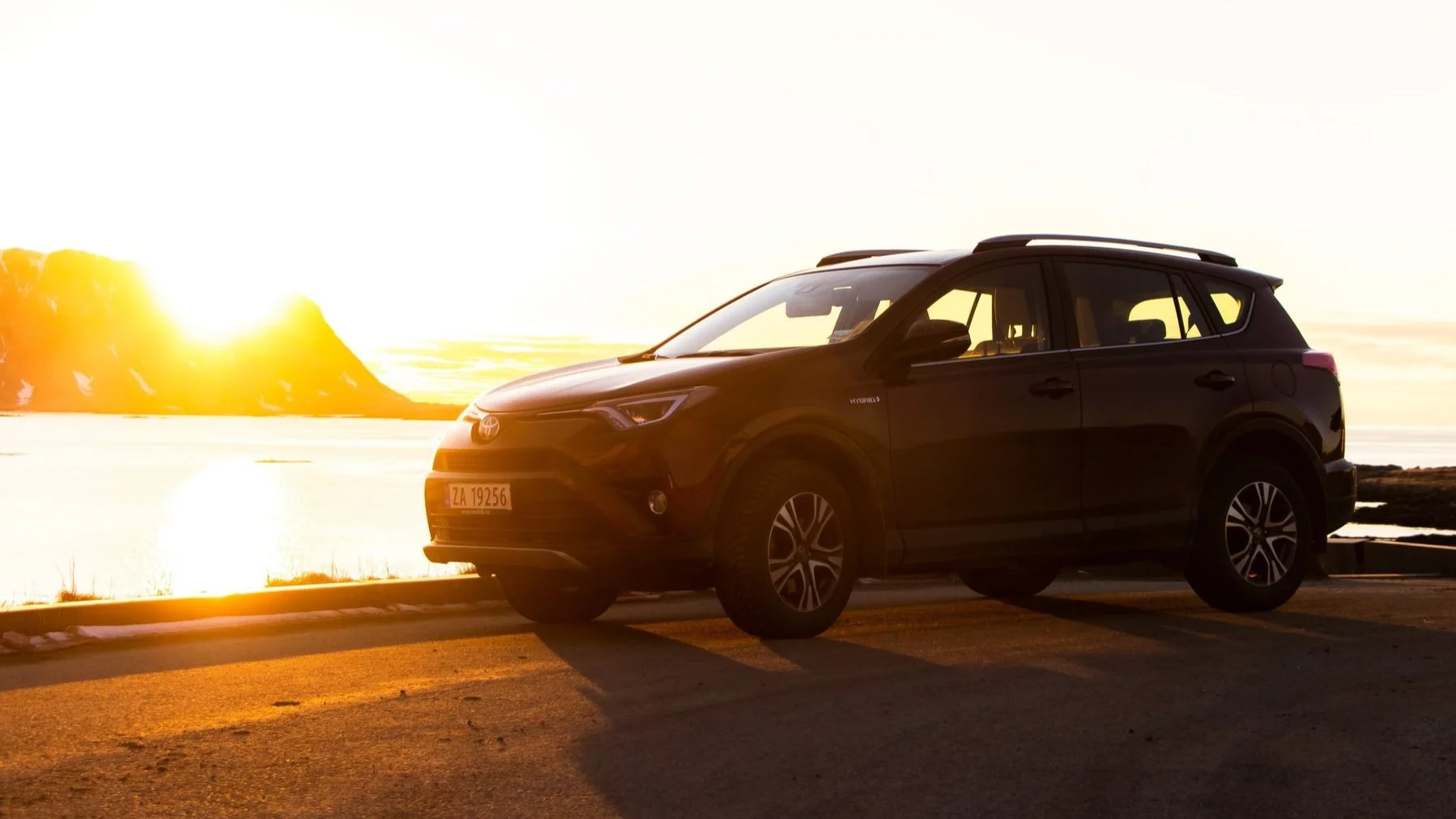 A black Toyota RAV4 hybrid parked by the water during sunset, with rocky cliffs in the background.