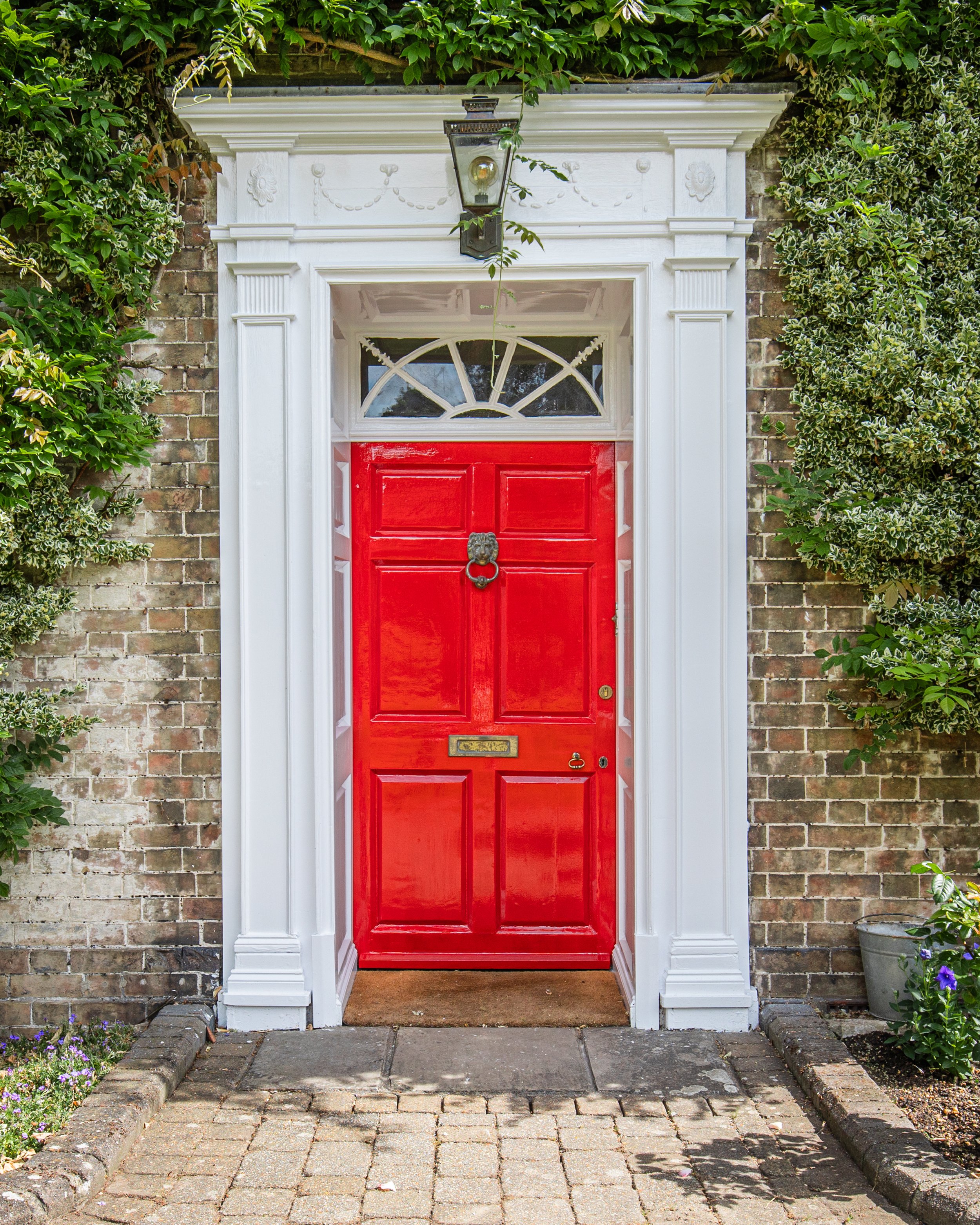 Red front door with decorative white trim and an arched window, surrounded by brick wall and greenery.
