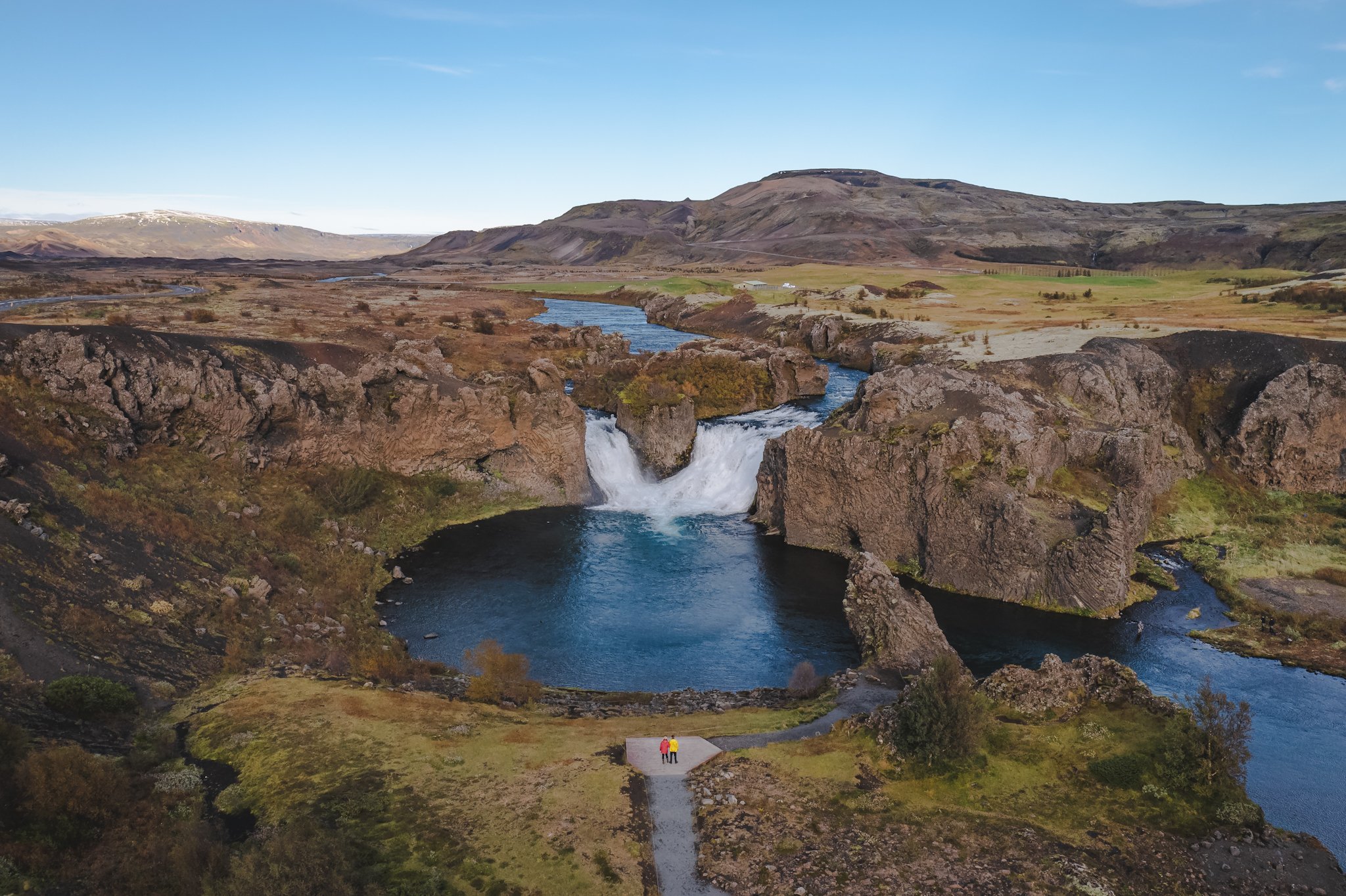 Two people standing on a small platform overlooking a river waterfall in a vast, rocky canyon. The landscape includes distant mountains, green fields, and a clear blue sky.