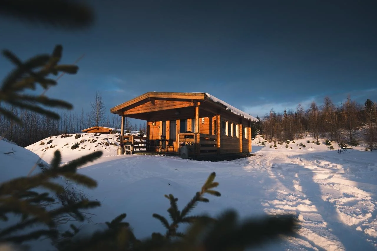 A wooden cabin in a snow-covered landscape during sunset, with a small porch and a distant wooded area.