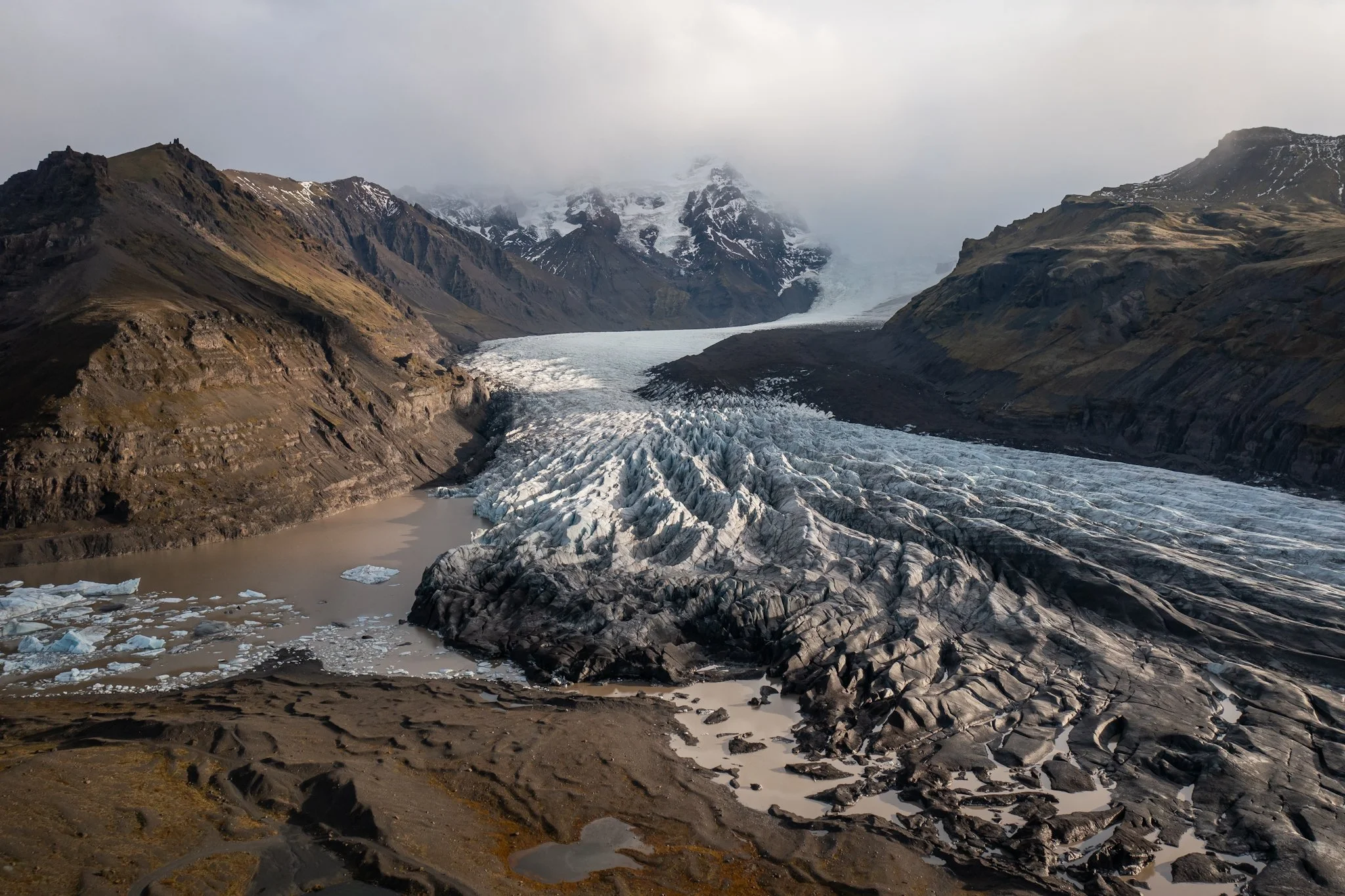 A glacier flowing between rugged mountains with snow patches and a cloudy sky.