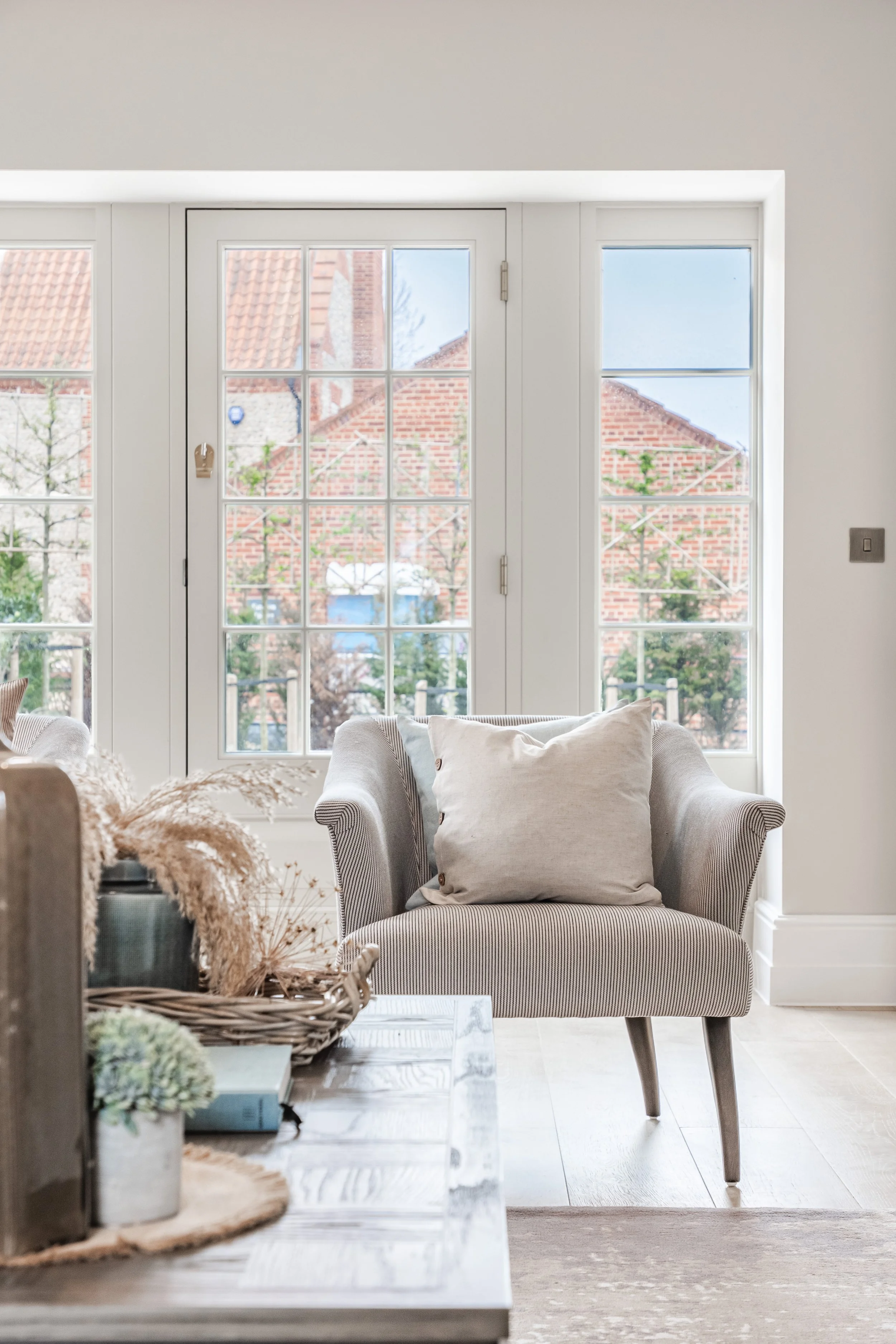 Living room with a beige armchair with a cream pillow, a table with a basket of dried flowers, and a large window showing houses and trees outside.