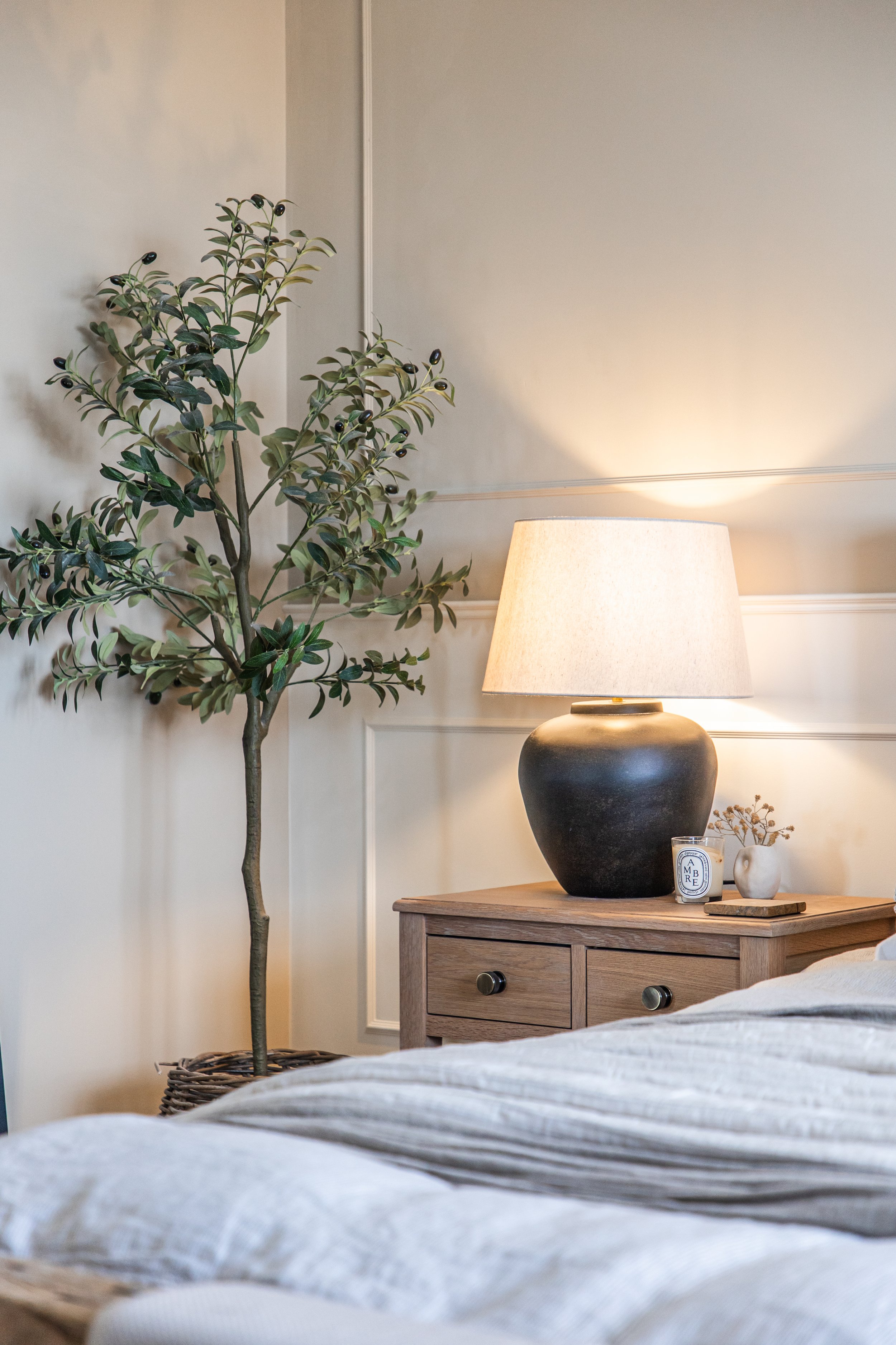 A cozy bedroom corner with a tall potted plant with dark berries on the left, a wooden nightstand with a large black vase, a lit table lamp, a candle, and a small white decorative object. The wall has decorative paneling and a soft light creates a wa