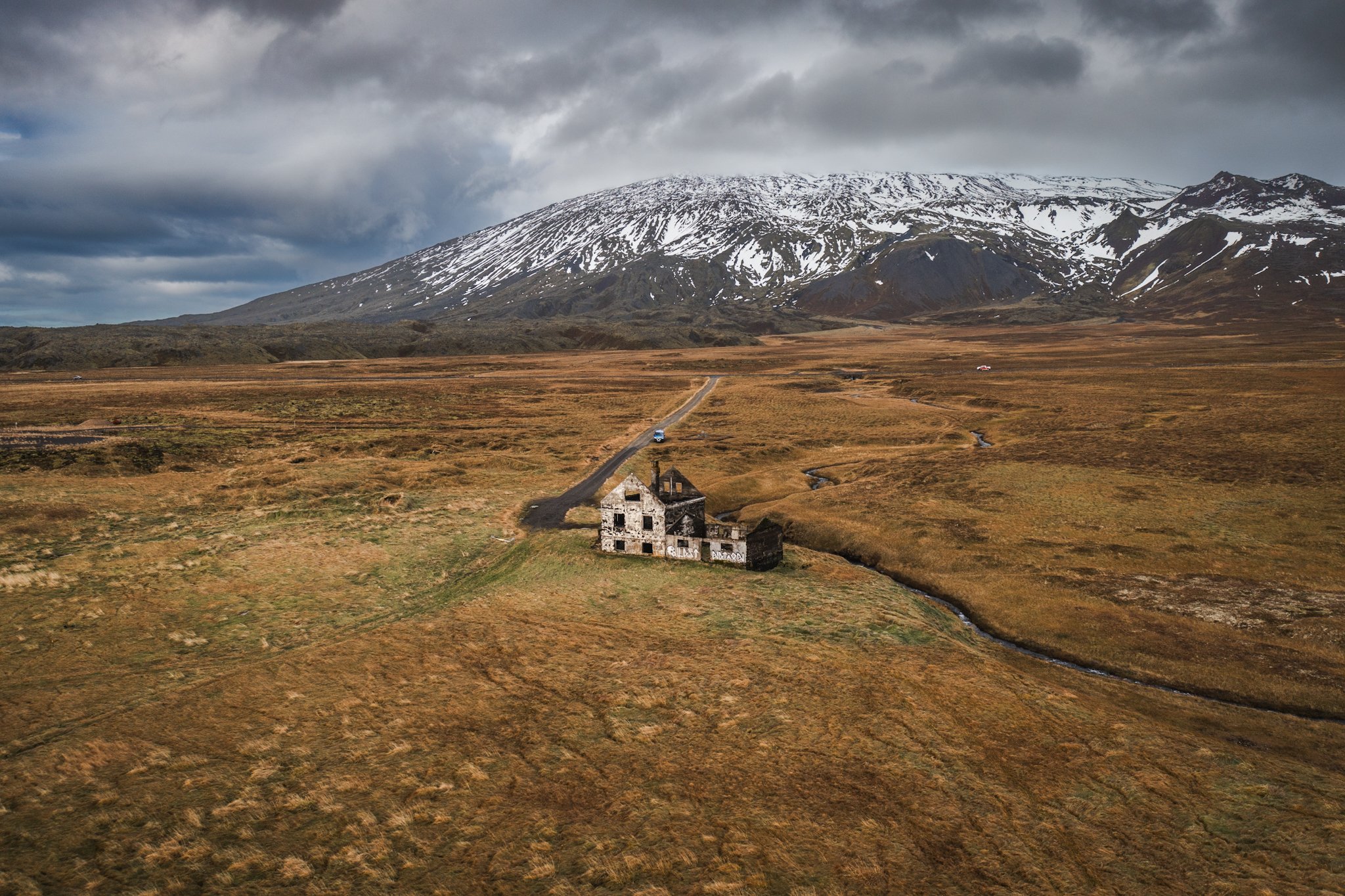 Remote landscape featuring an abandoned, dilapidated house with graffiti, a winding dirt road, open grasslands, and a snow-capped mountain under cloudy sky.