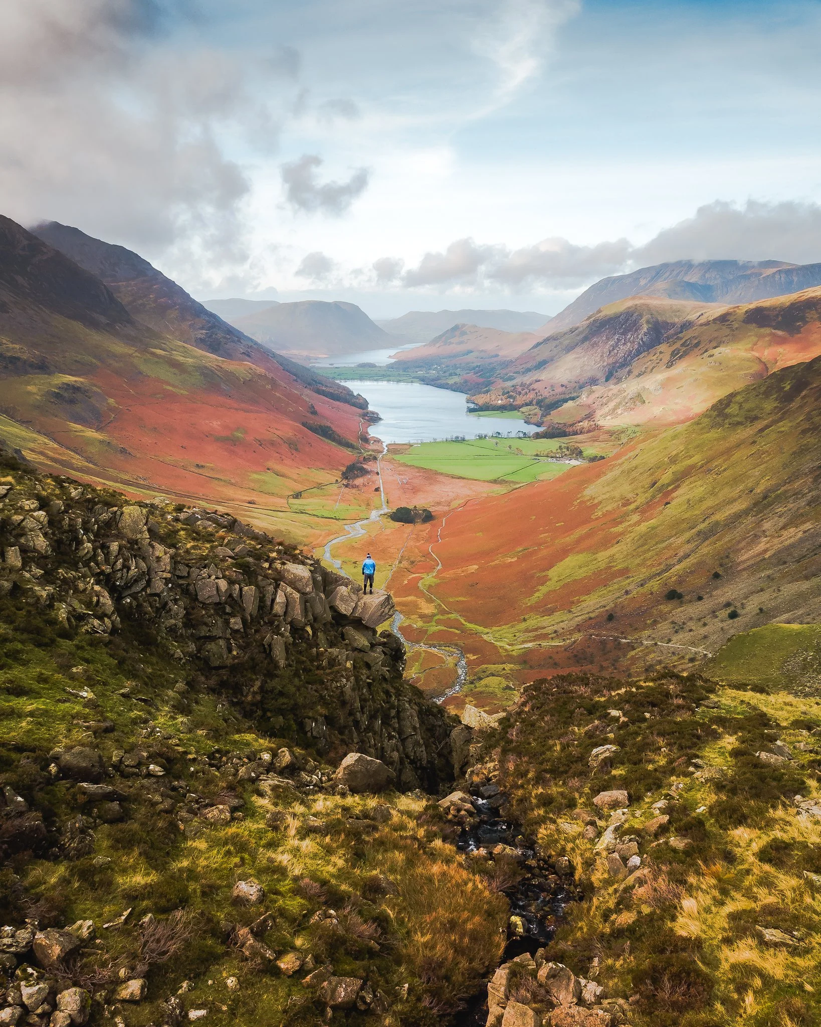 A scenic view of a river flowing through a valley surrounded by mountains with a cloudy sky above.