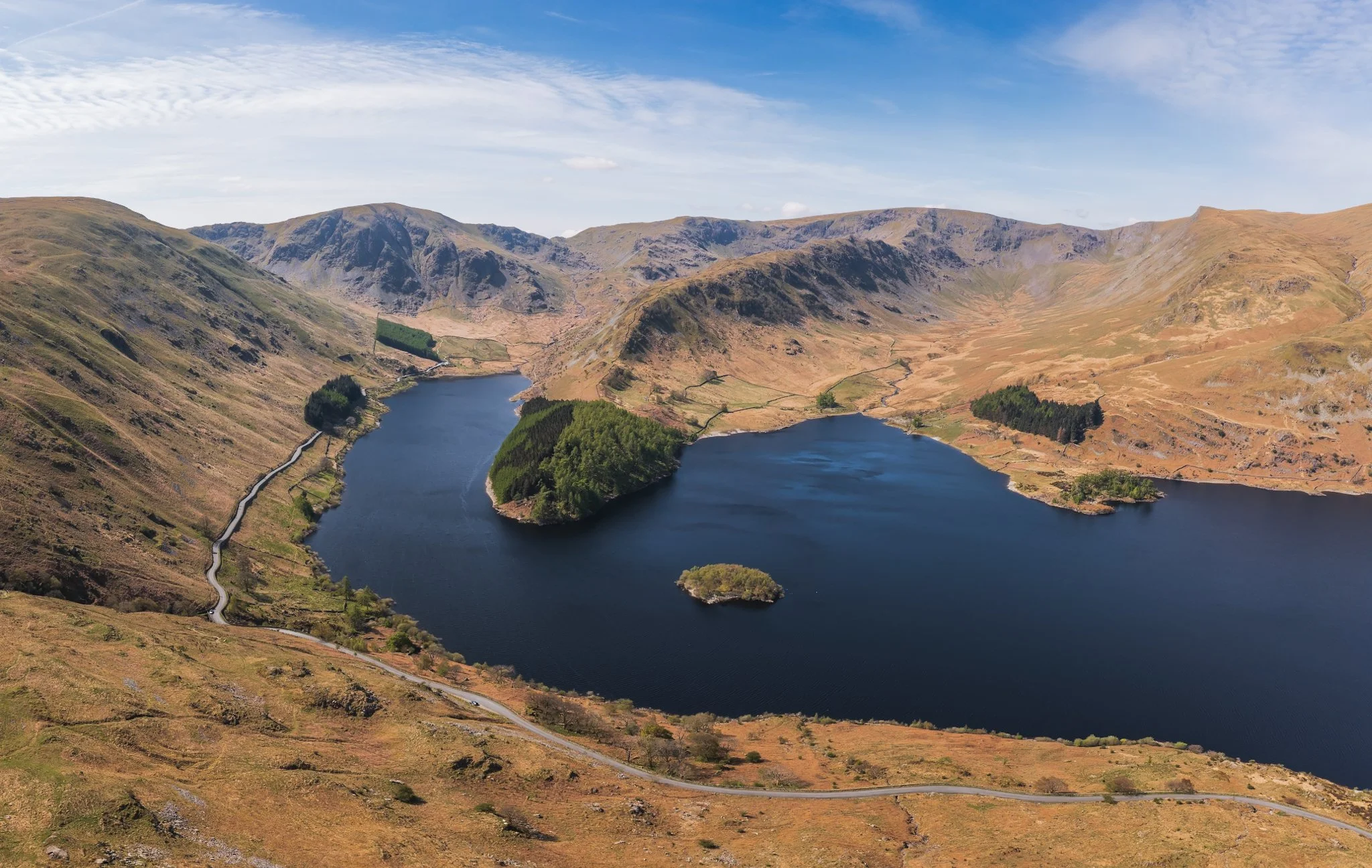 Aerial view of a mountain landscape featuring a deep blue lake surrounded by rolling hills with patches of greenery and a winding road.
