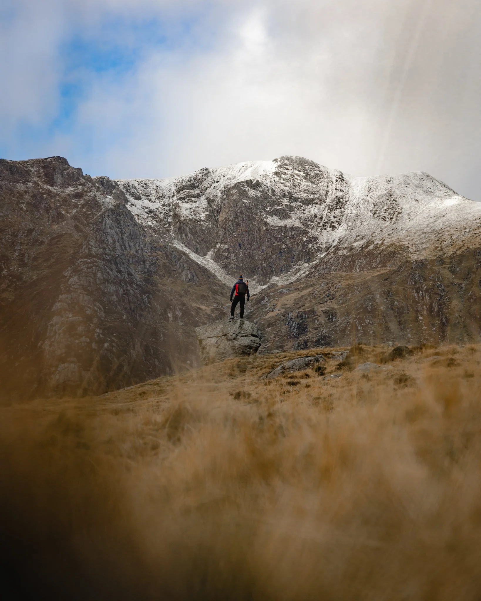 A hiker standing on rocky terrain with snow-capped mountains in the background, under a cloudy sky.