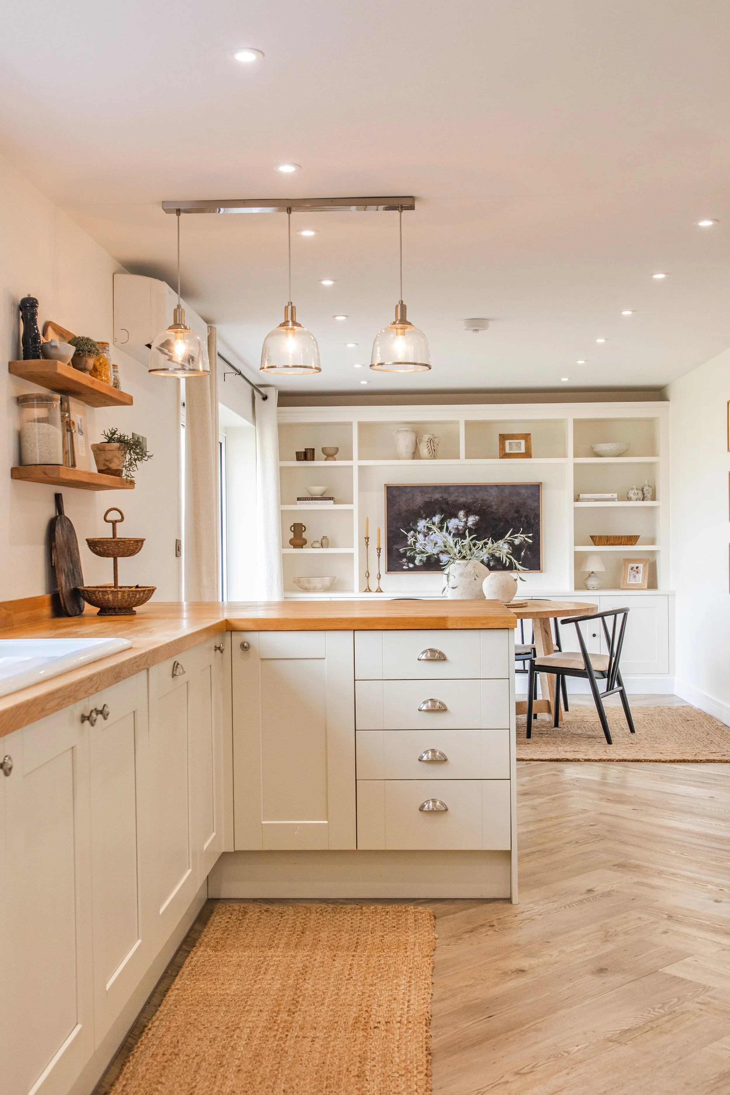 A cozy, modern kitchen and dining area with white cabinetry, wooden countertops, decorative shelving, pendant lights, and an open shelving wall with centered artwork and decorative items. A hallway with a rug leads to the dining space with chairs and