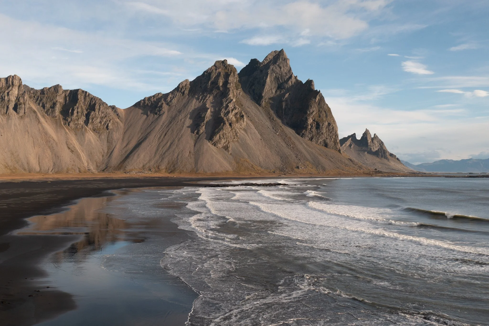 A rugged mountain range next to the ocean with waves crashing onto a dark sand beach.